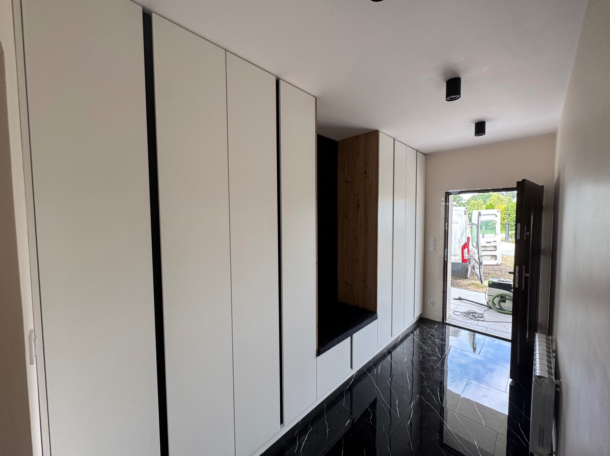 Biała szafa wnękowa z siedziskiem z ciemnego drewna i czarną tapicerką, na tle błyszczącej, czarnej podłogi. Widok na korytarz i otwarte drzwi wejściowe.