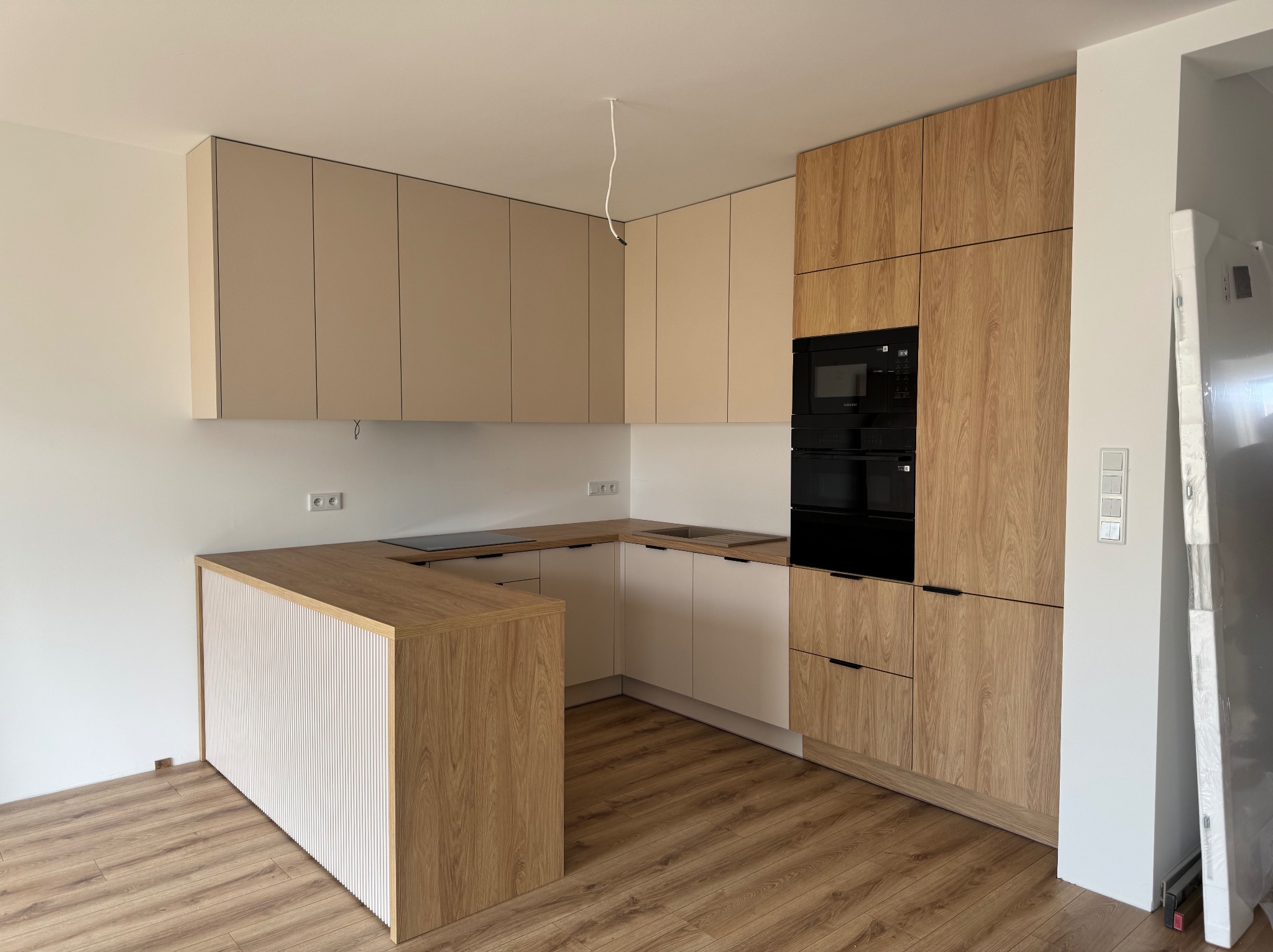 Nowoczesna kuchnia w kształcie litery U z drewnianymi i beżowymi frontami, czarnym piekarnikiem i płytą indukcyjną. Widoczne detale wykończenia i oświetlenie.