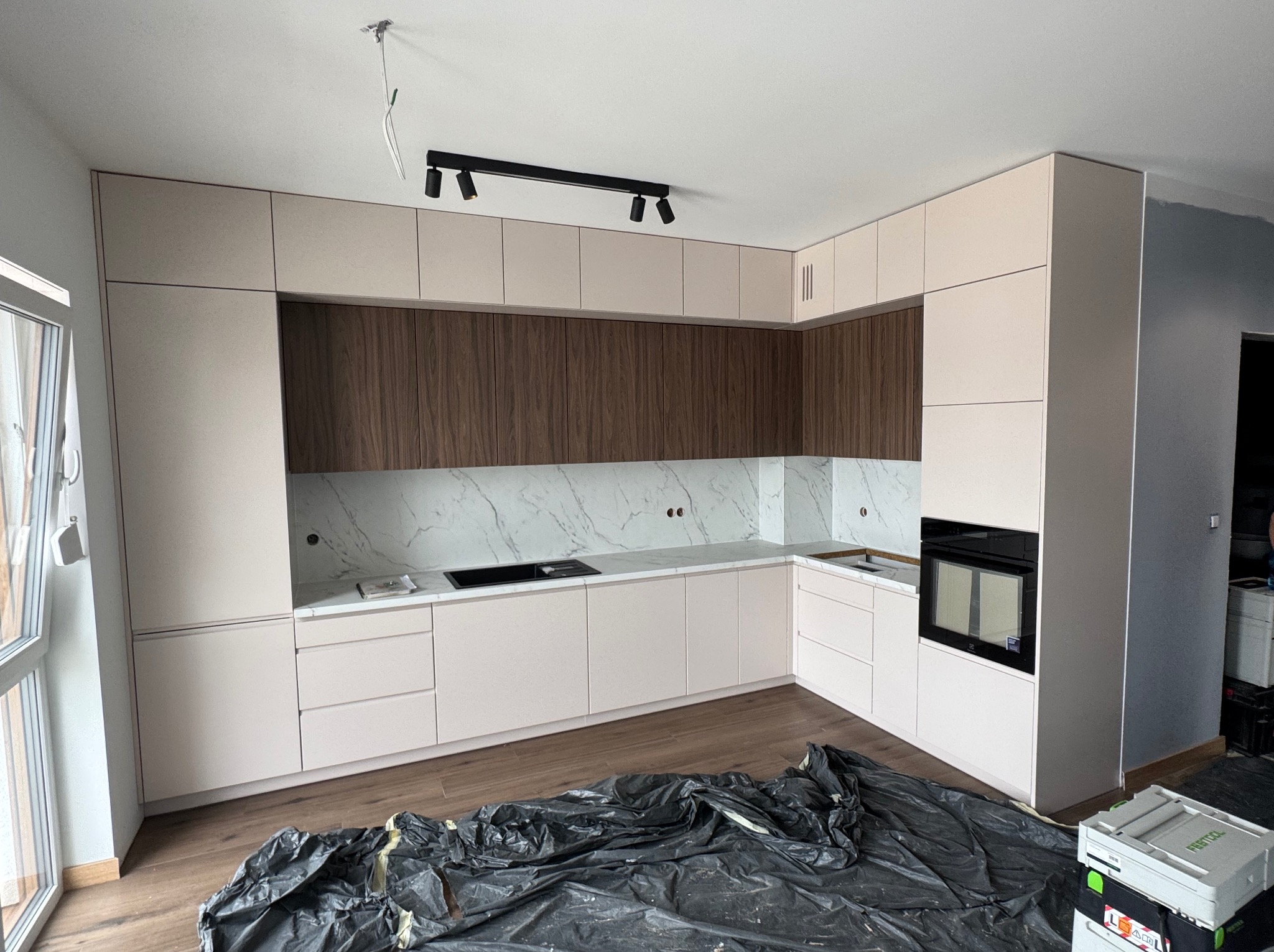 Jasna kuchnia na wymiar z drewnianymi akcentami i marmurowym blatem, widok z perspektywy podłogi, zabezpieczonej folią malarską. Widoczne narzędzia.