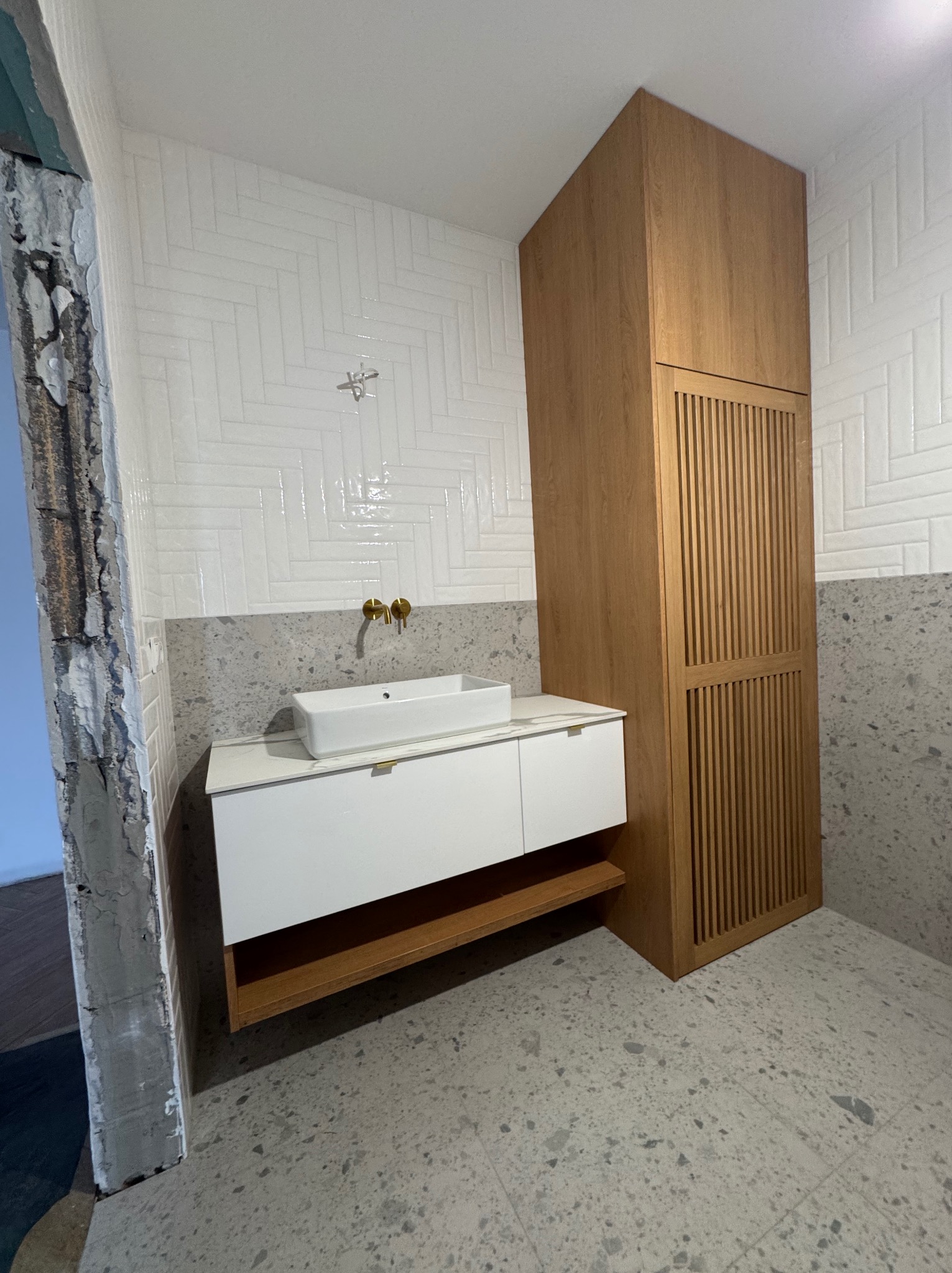 Nowoczesna łazienka z białą umywalką nablatową, szafką z drewnianym akcentem i wysoką szafą. Ściany wyłożone białymi płytkami w jodełkę i szarym lastryko.