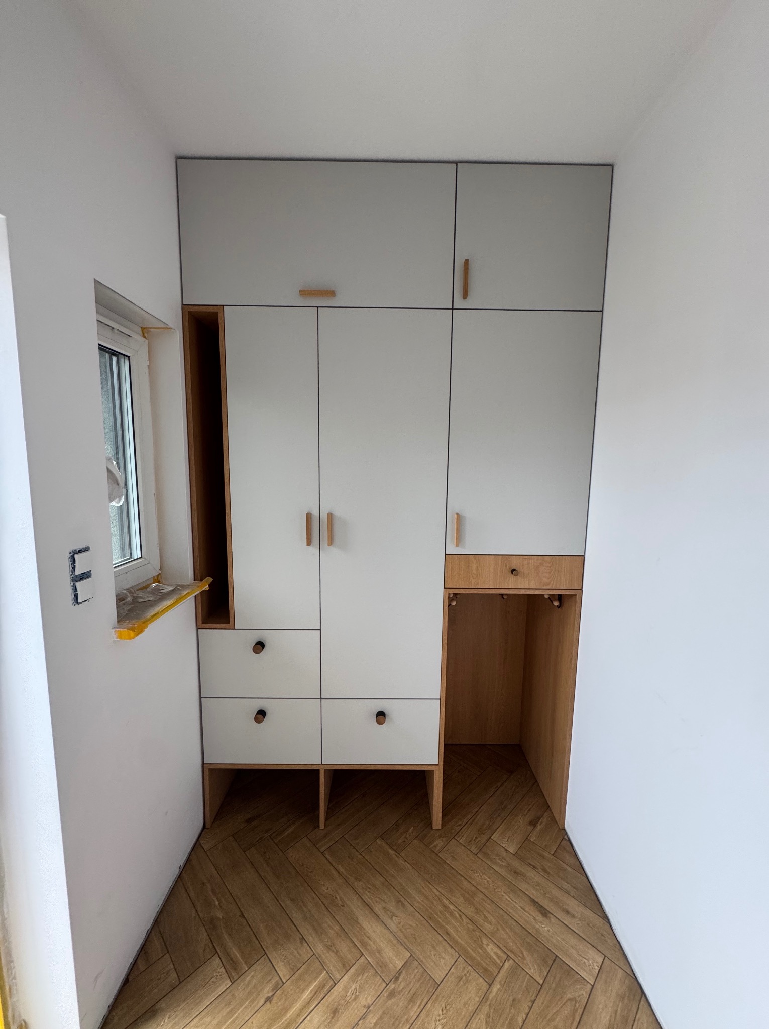 Jasnoszara szafa wnękowa z drewnianymi akcentami, wkomponowana w przestrzeń z oknem i parkietem w jodełkę. Minimalistyczny design, naturalne materiały.