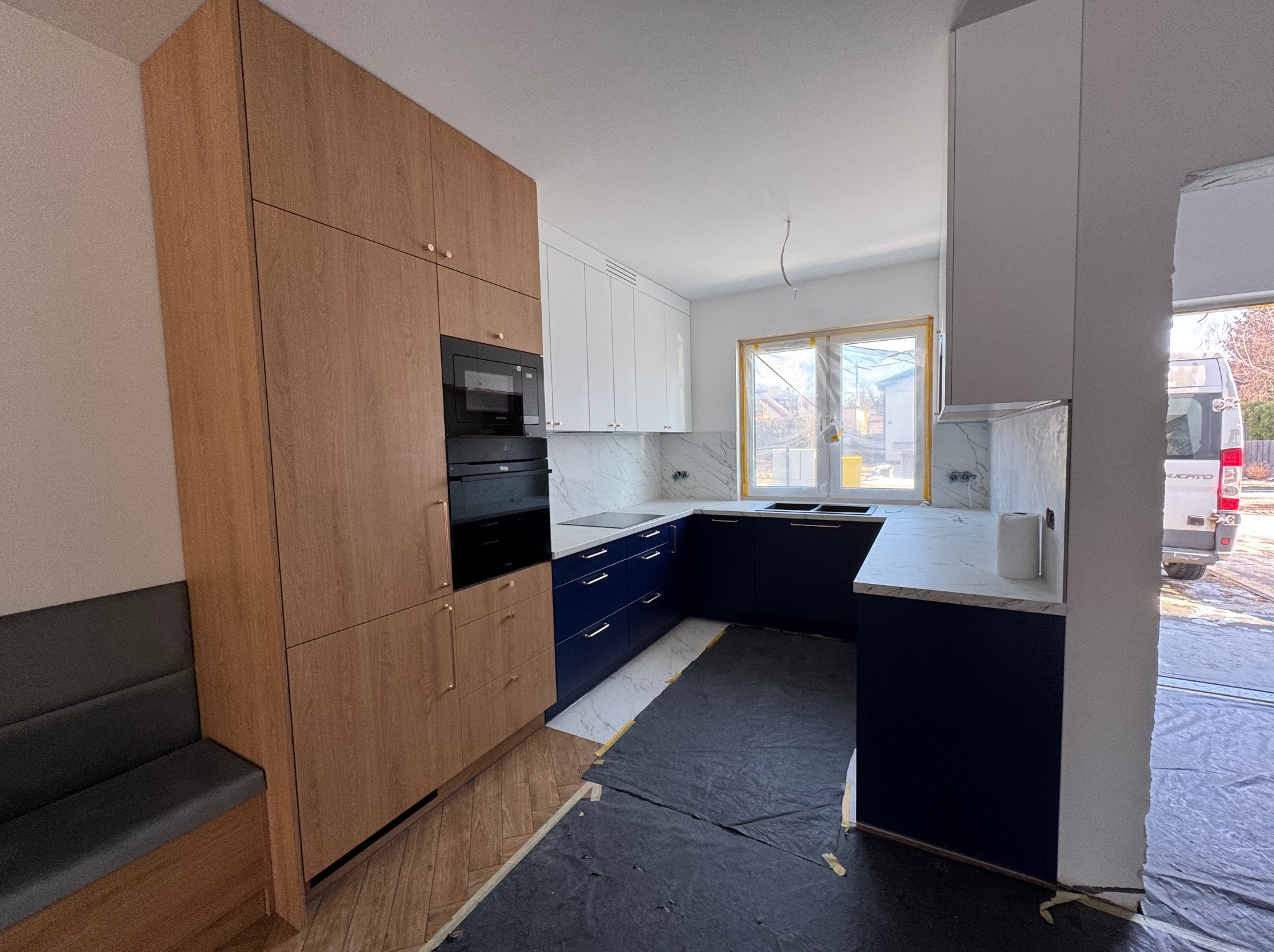 Nowoczesna kuchnia w trakcie montażu: granatowe szafki dolne, białe górne, drewnopodobne wysokie zabudowy z piekarnikiem, marmurowy blat. Podłoga zabezpieczona folią.