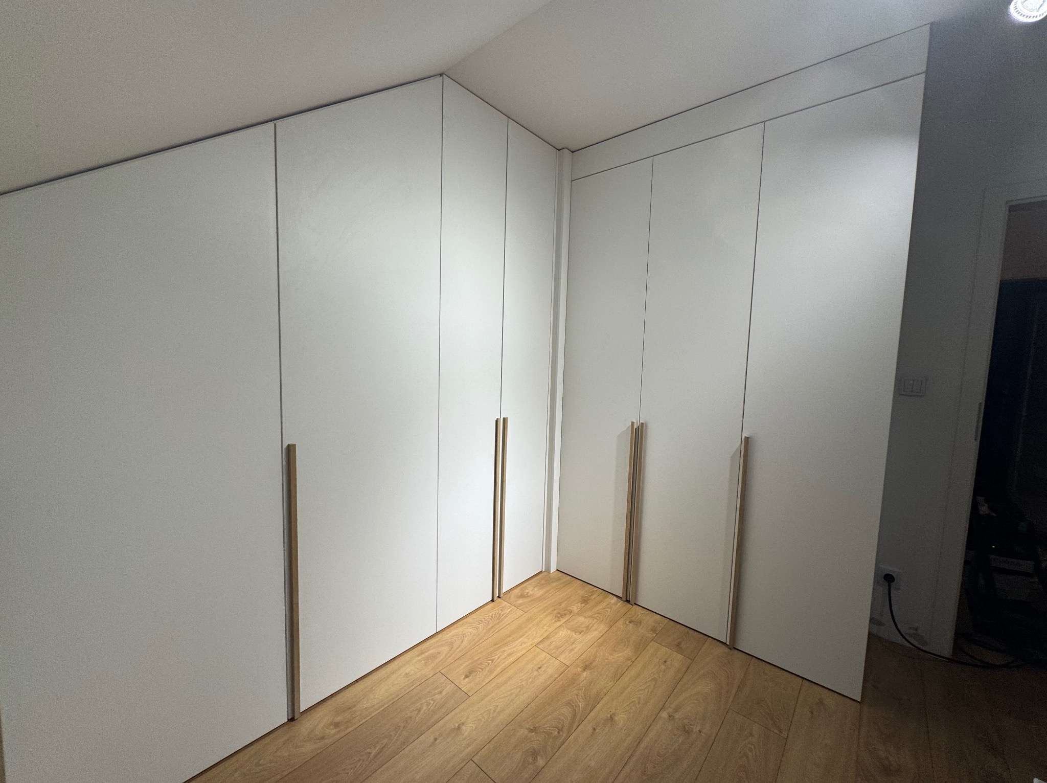 Biała szafa wnękowa z drewnianymi uchwytami, dopasowana do skosu pomieszczenia, na podłodze panele drewniane. Widok na narożnik szafy.