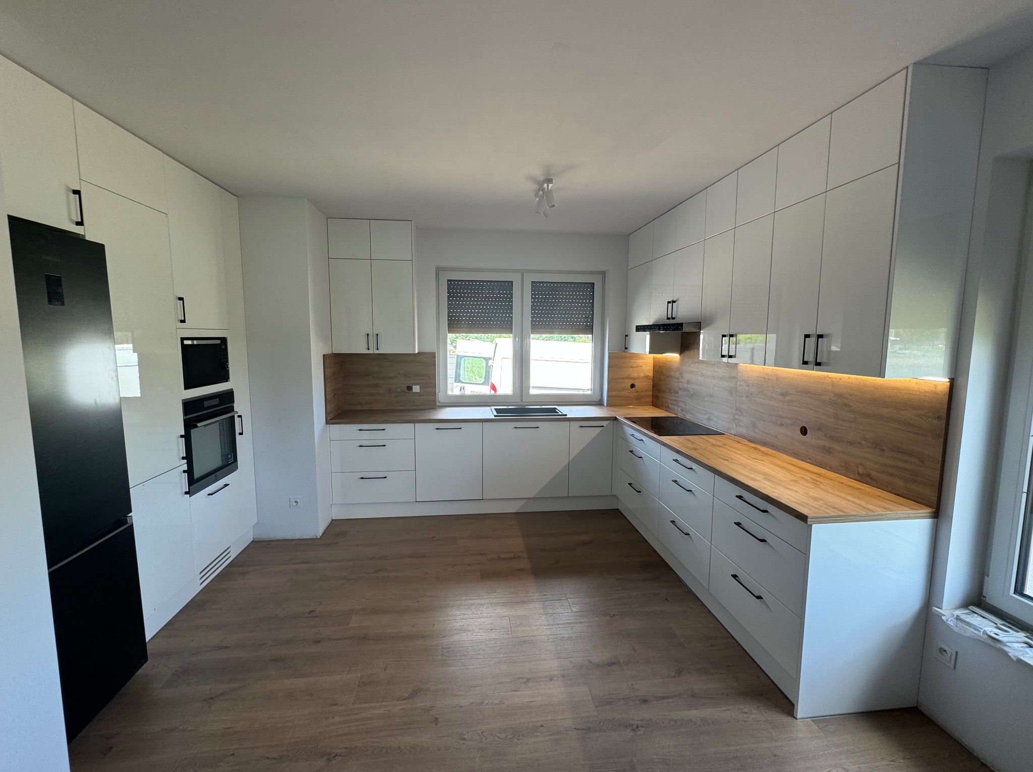 Nowoczesna, jasna kuchnia z białymi, lakierowanymi frontami i drewnianym blatem. Czarny sprzęt AGD. Minimalistyczne uchwyty. Widok na okno z roletami.