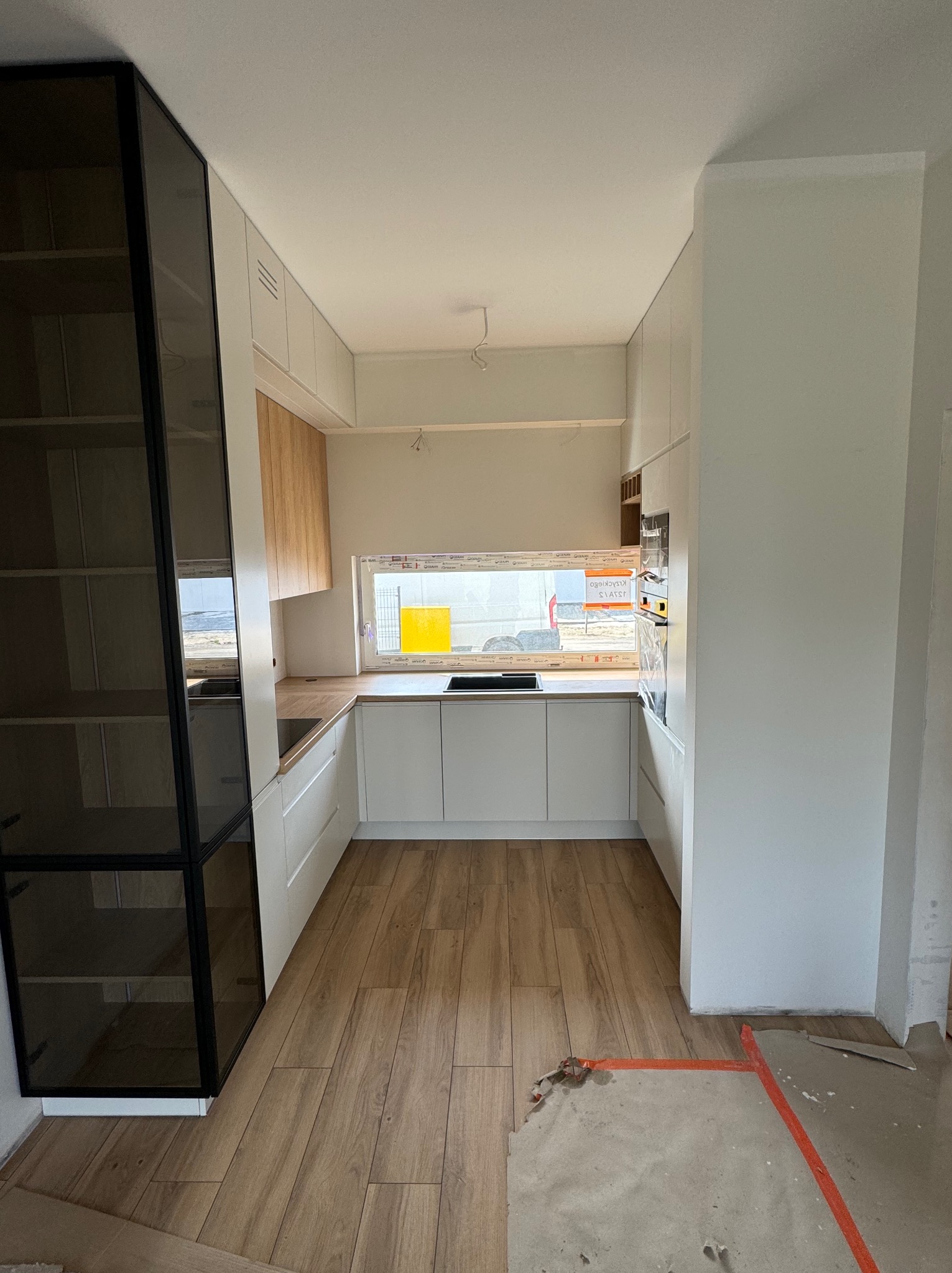 Nowoczesna kuchnia z białymi szafkami i drewnianymi akcentami, widok na okno z żółtą folią ochronną, drewniana podłoga, częściowo zakryta folią malarską.
