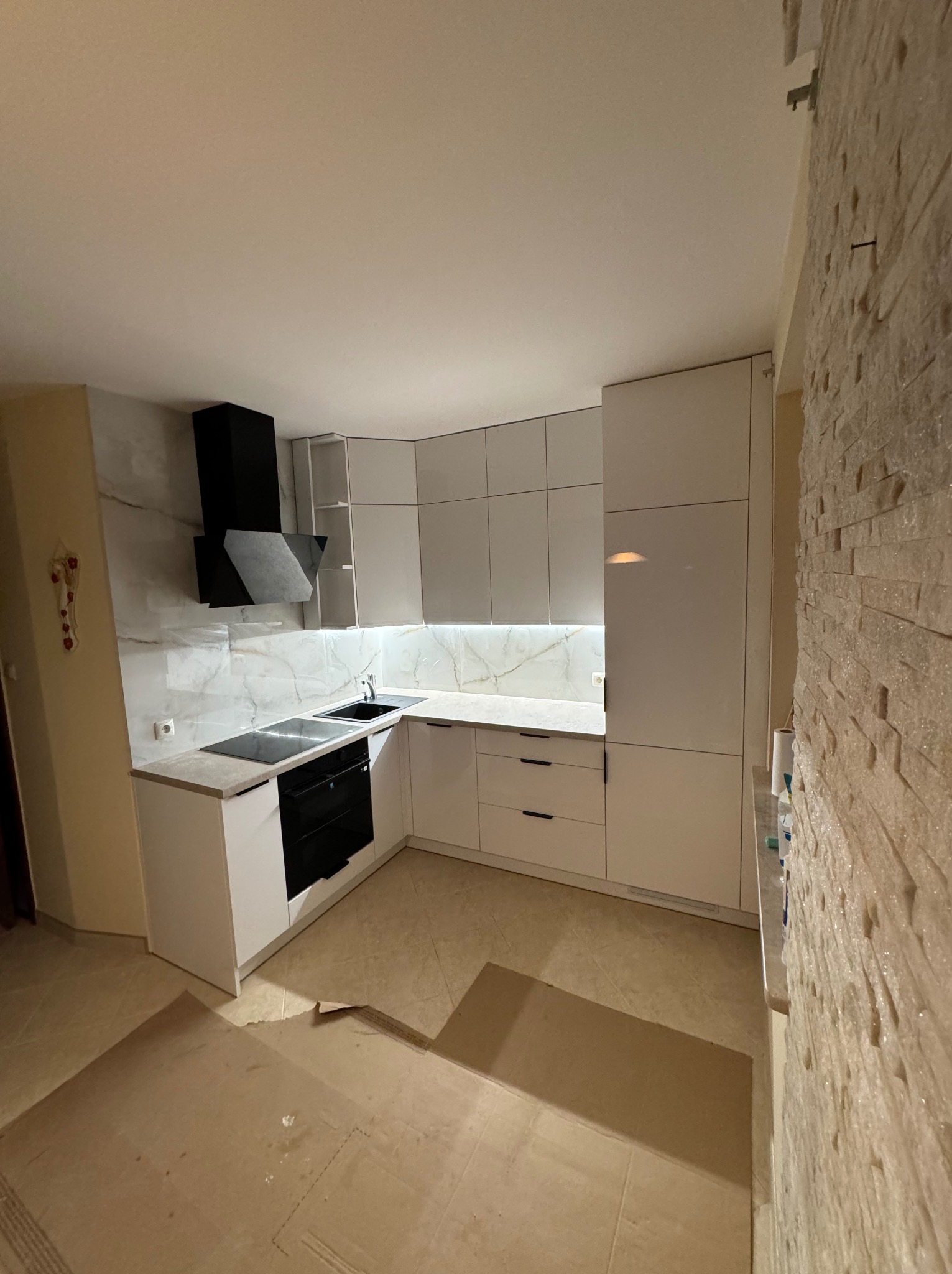 Nowoczesna, biała kuchnia z czarnym okapem i marmurowym backsplashiem. Uwagę przykuwa wykończenie ściany z dekoracyjnej cegły i podświetlenie LED pod szafkami.