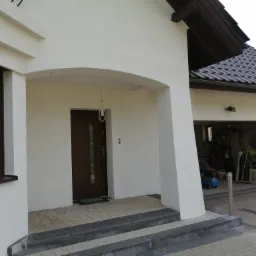 Biała elewacja domu jednorodzinnego z drewnianą stolarką okienną i drzwiami, widoczny podjazd z kostki brukowej i otwarty garaż z wyposażeniem.