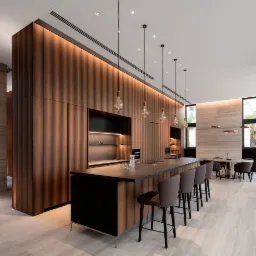 Nowoczesna kuchnia z wyspą, drewniane fronty szafek z podświetleniem LED, szare krzesła barowe, marmurowe płytki podłogowe i minimalistyczne lampy wiszące.