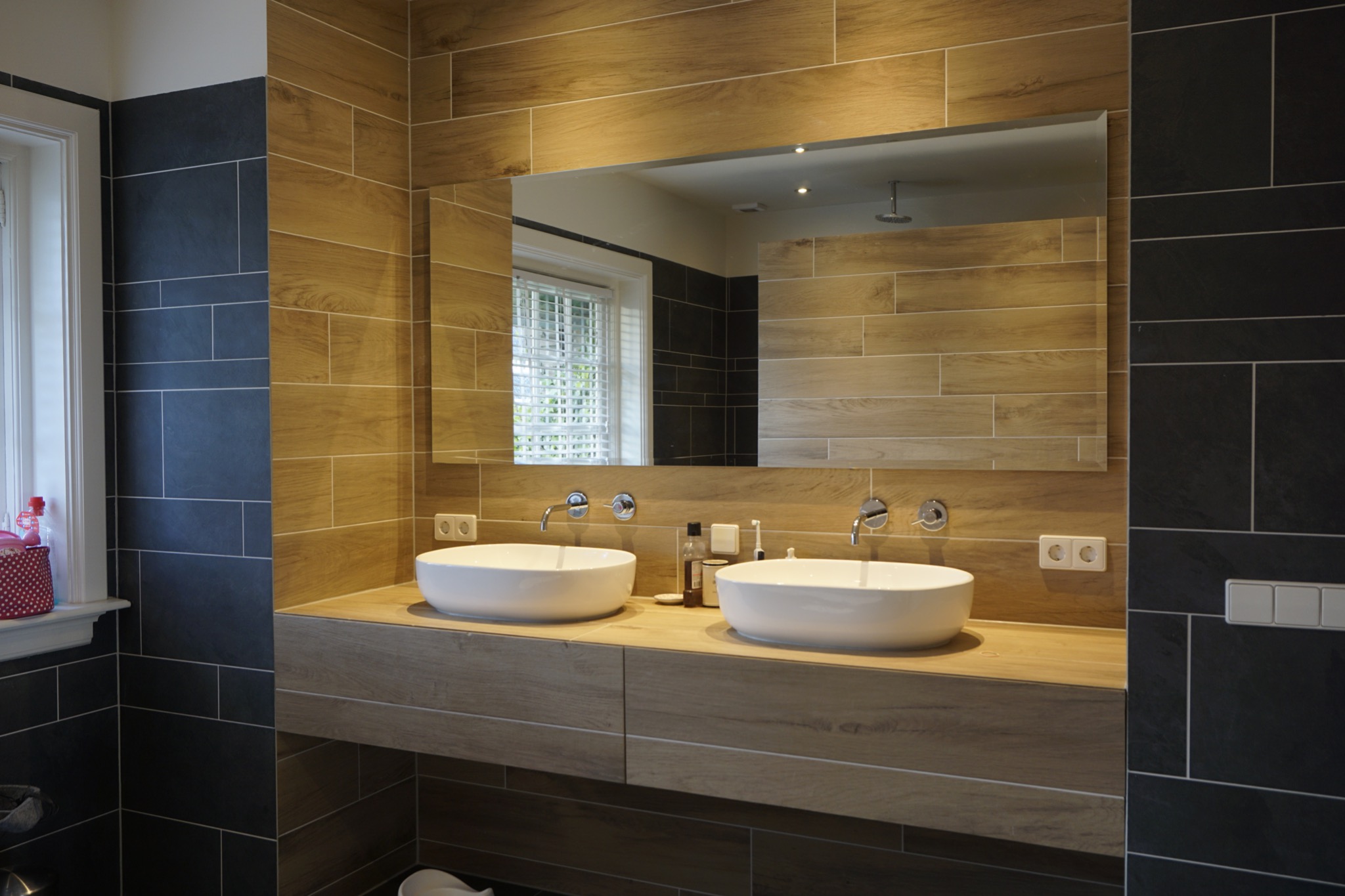 Nowoczesna łazienka z dwoma okrągłymi umywalkami nablatowymi, drewnianą zabudową szafek pod umywalkami i lustrem bez ramy, drewnopodobne płytki na ścianach zestawione z ciemnymi płytkami.