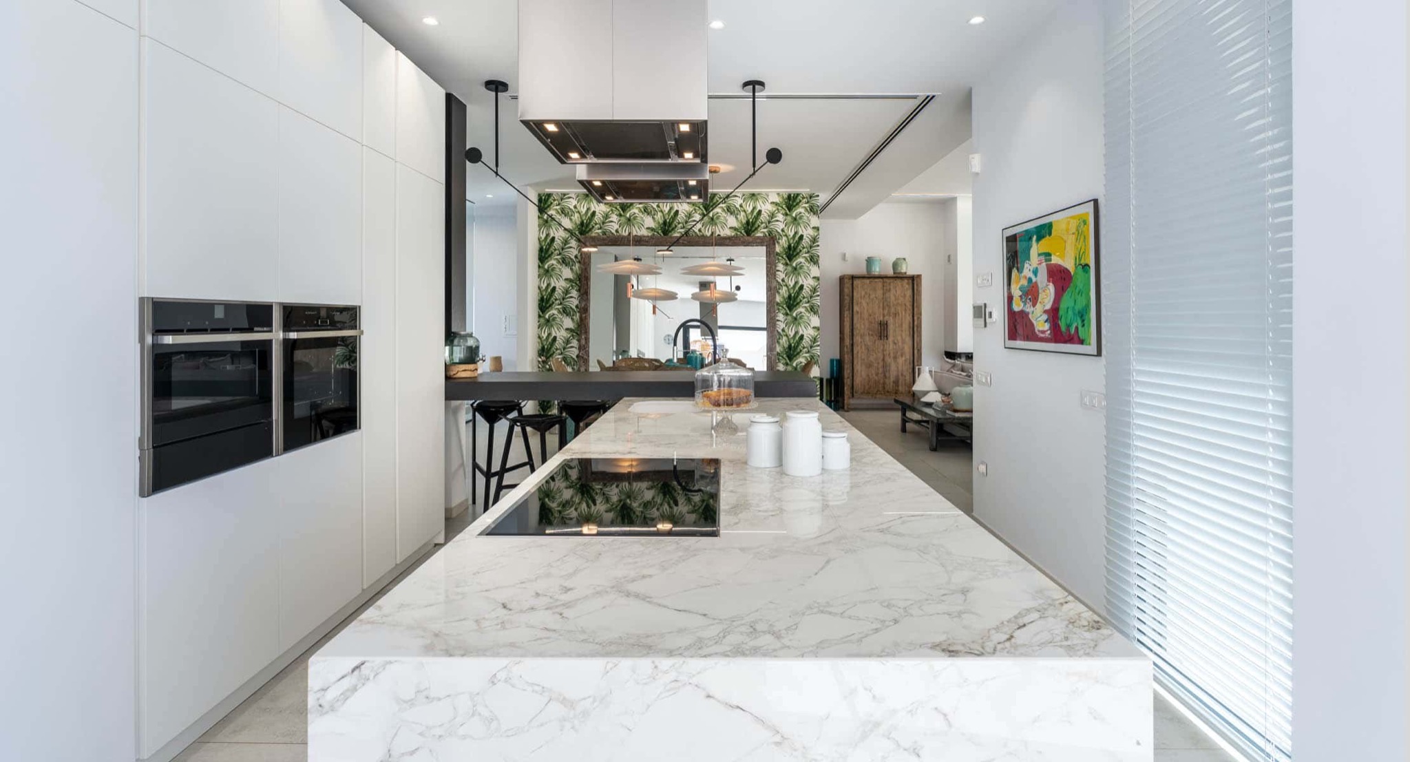 Nowoczesna kuchnia z marmurowym blatem wyspy, wbudowaną płytą grzewczą i białymi, minimalistycznymi szafkami, oświetlona wiszącymi lampami i z lustrem z motywem tropikalnym w tle.