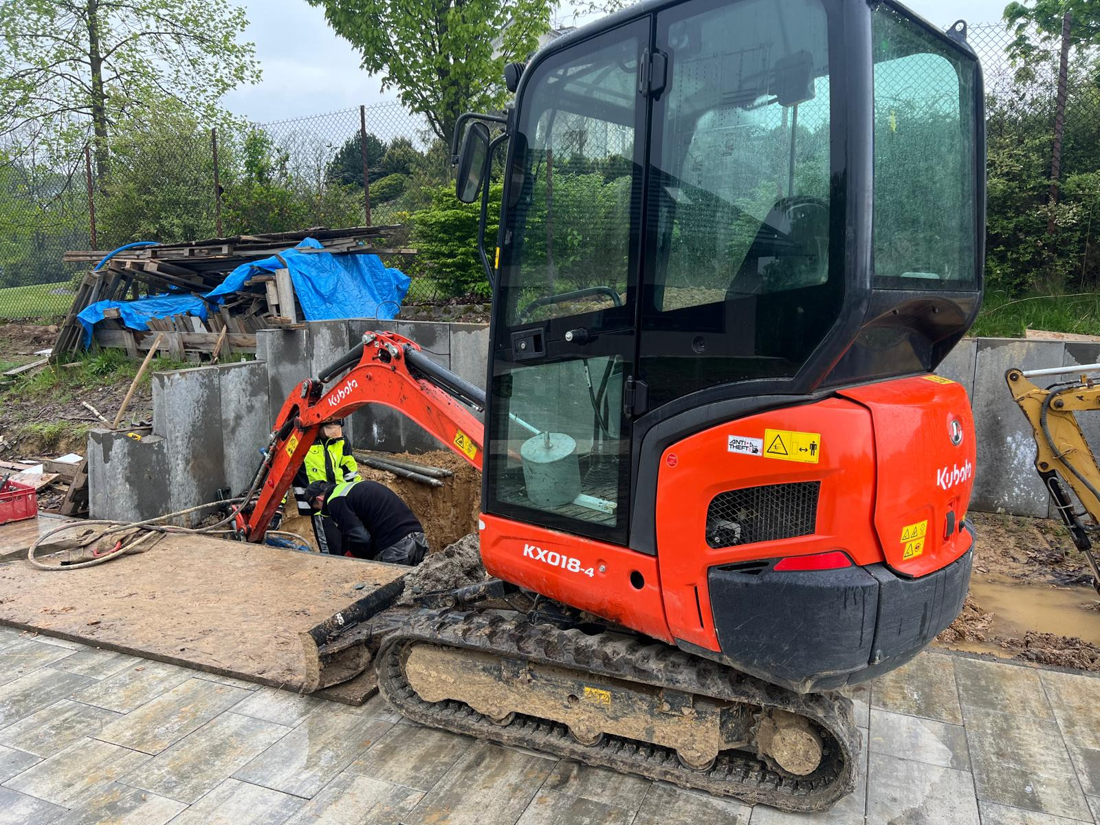 Pomarańczowa mini koparka Kubota KX018-4 na błotnistym terenie z pracownikiem w żółtej kamizelce w wykopie, widoczne betonowe ściany wykopu i zaplecze budowy z paletami i plandeką.