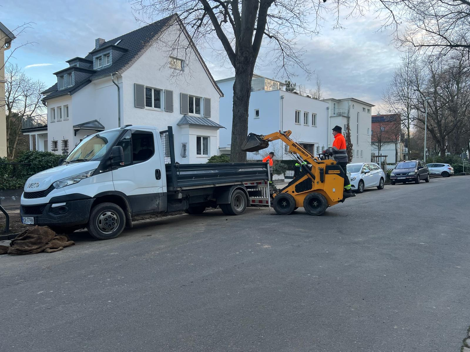 Żółta miniładowarka w Krakowie załadowuje ziemię na skrzynię białego samochodu dostawczego na tle zabudowy miejskiej.