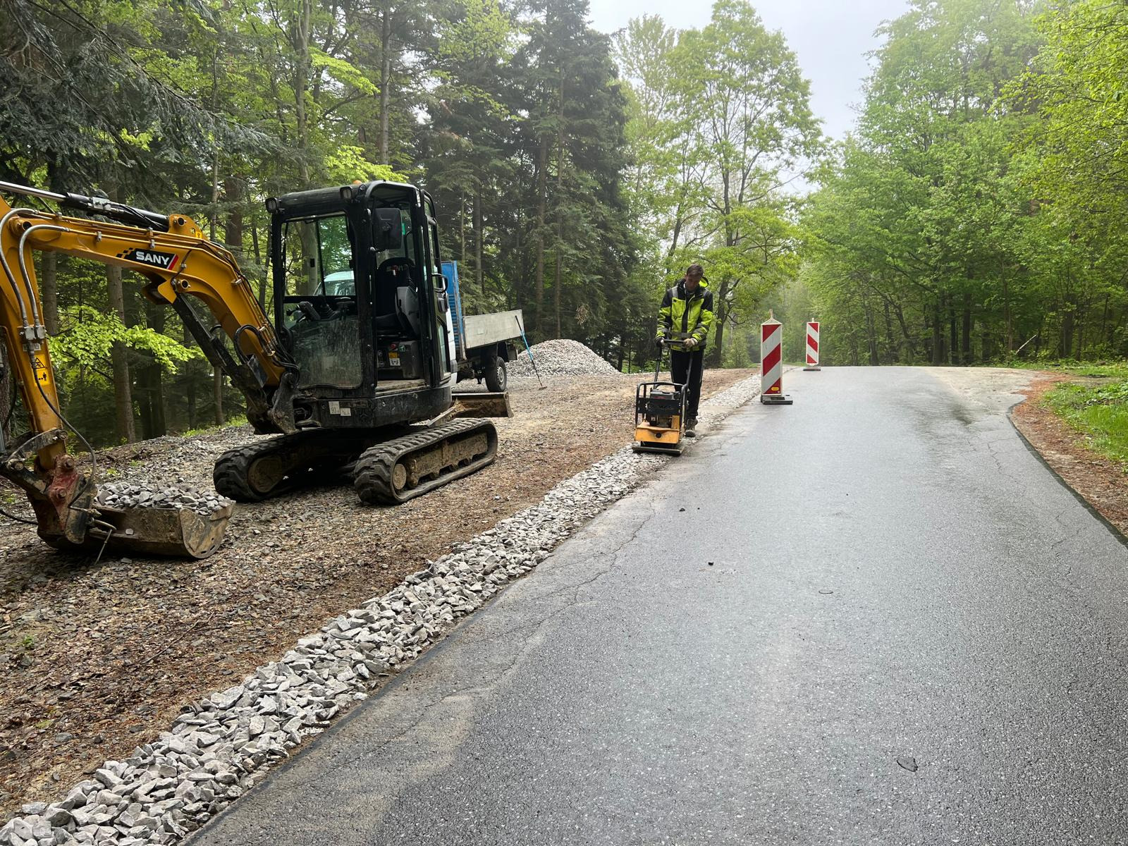 Czarny mini-koparka SANY i pracownik w jaskrawej kamizelce zagęszczający podłoże ubijarką wibracyjną na obrzeżach asfaltowej drogi, obok pryzmy kamieni i słupków ostrzegawczych w otoczeniu leśnym.