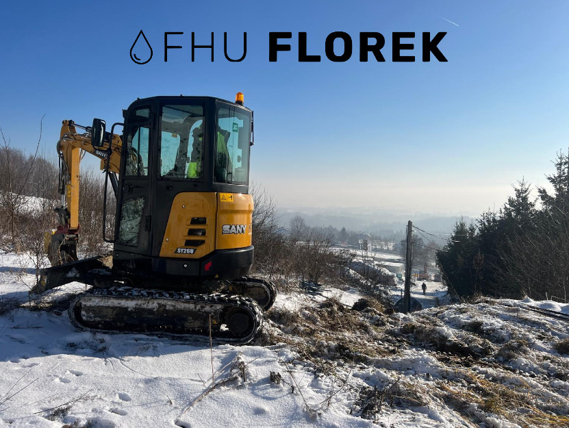Żółta mini koparka SANY SY26U na zaśnieżonym terenie, widoczne ślady pracy, w tle panorama miasta pod błękitnym niebem, logo firmy FHU Florek u góry.