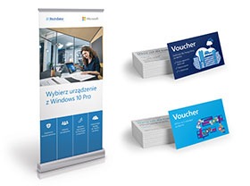 Roll-up reklamowy z hasłem 'Wybierz urządzenie z Windows 10 Pro', wizytówki ułożone w stosy oraz dwie pojedyncze wizytówki z motywem voucherów w kolorach niebieskim i białym.