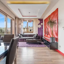 Nowoczesny salon z dużym oknem balkonowym, skórzaną sofą, fioletowym dywanem i oryginalną dekoracją ścienną w kolorze czerwonym.