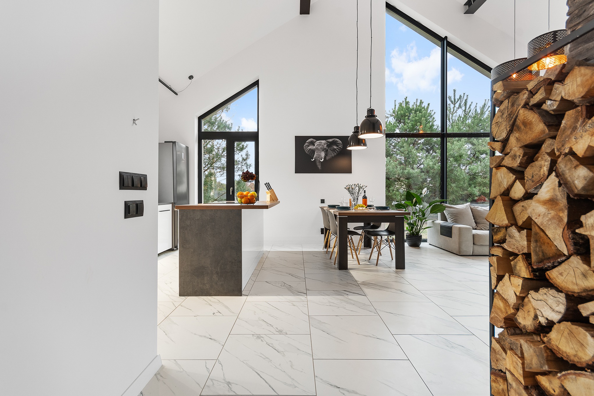 Jasne, przestronne wnętrze domu z wysokim sufitem, dużymi oknami, marmurową podłogą, wyspą kuchenną i stosem drewna obok salonu.