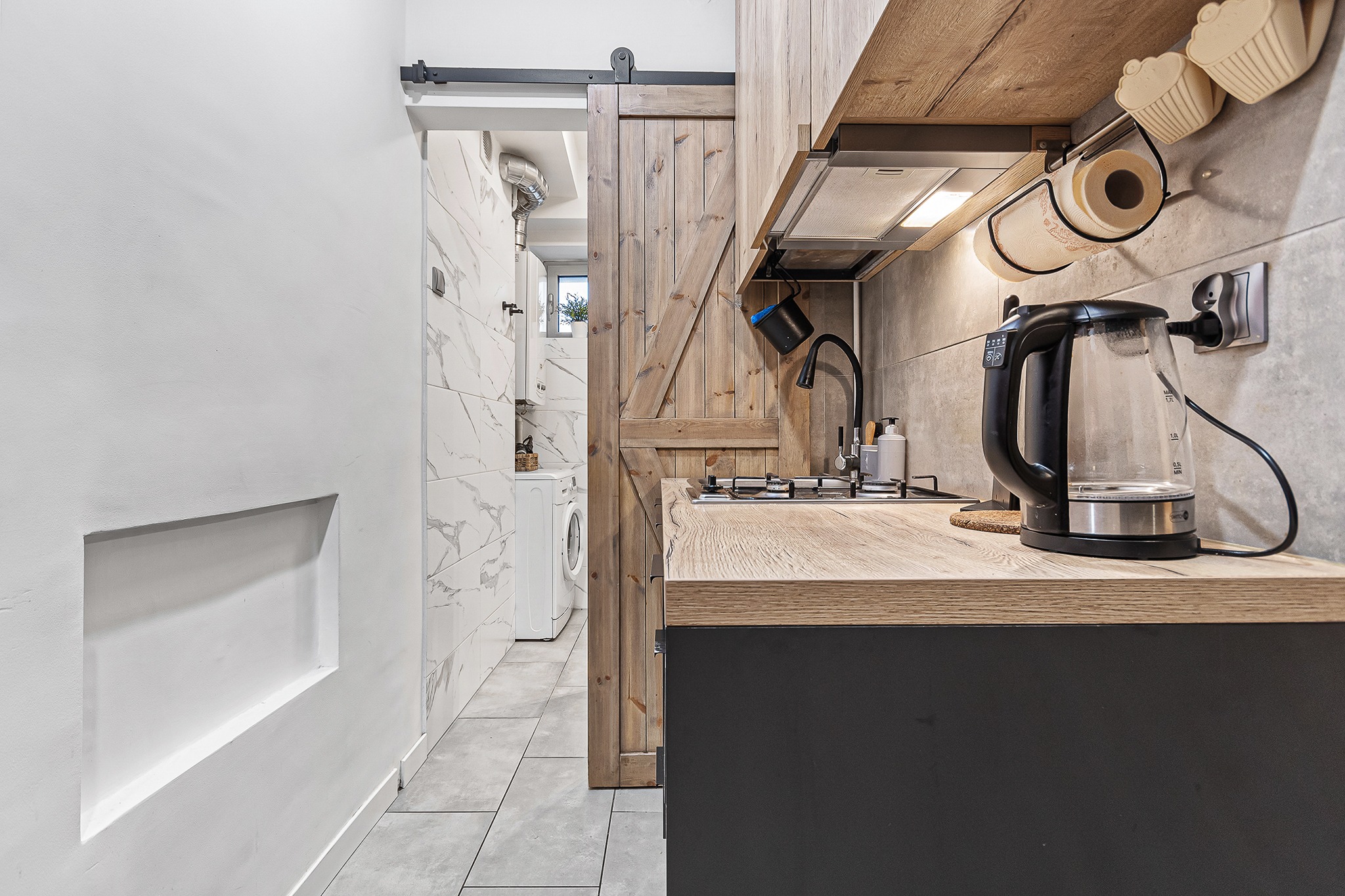 Wnętrze małej kuchni z drewnianą zabudową, przesuwnymi drzwiami stodołowymi i widokiem na pralnię z pralką.