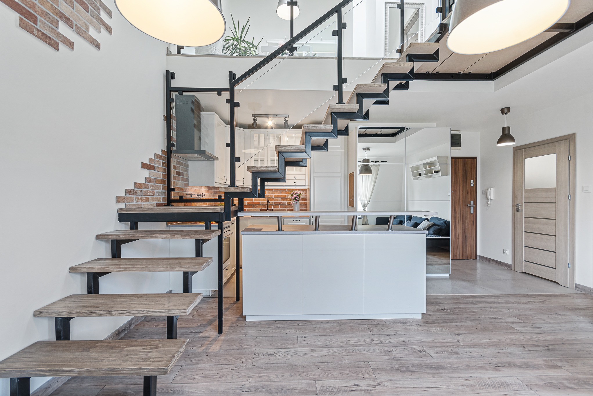 Dwupoziomowe mieszkanie z otwartą kuchnią, schodami z drewnianymi stopniami i czarną metalową konstrukcją oraz szklaną balustradą, widok na salon i drzwi wejściowe