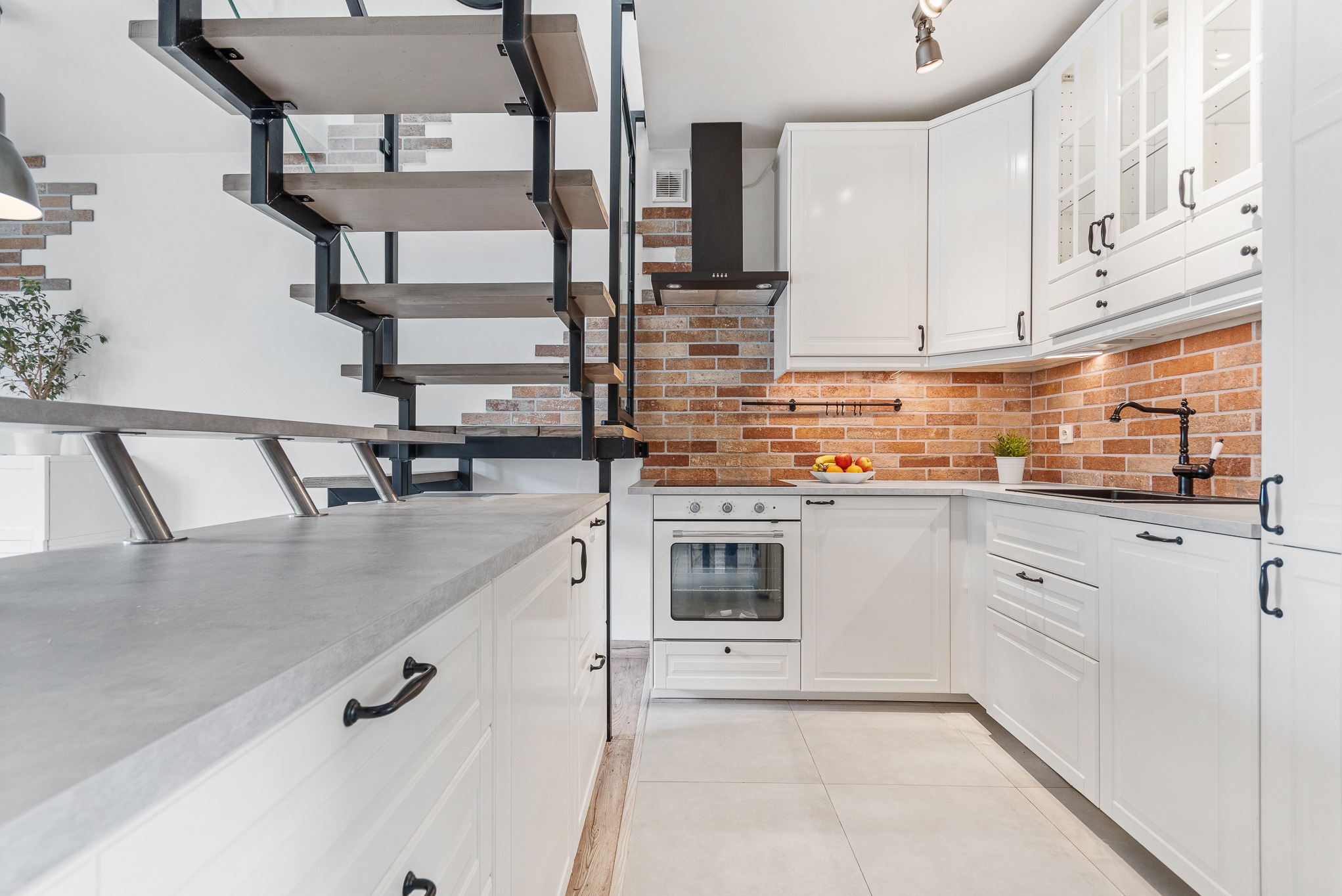 Nowoczesna, biała kuchnia z ceglaną ścianą i schodami z drewnianymi stopniami na metalowej konstrukcji, oświetlona punktowymi lampami sufitowymi.