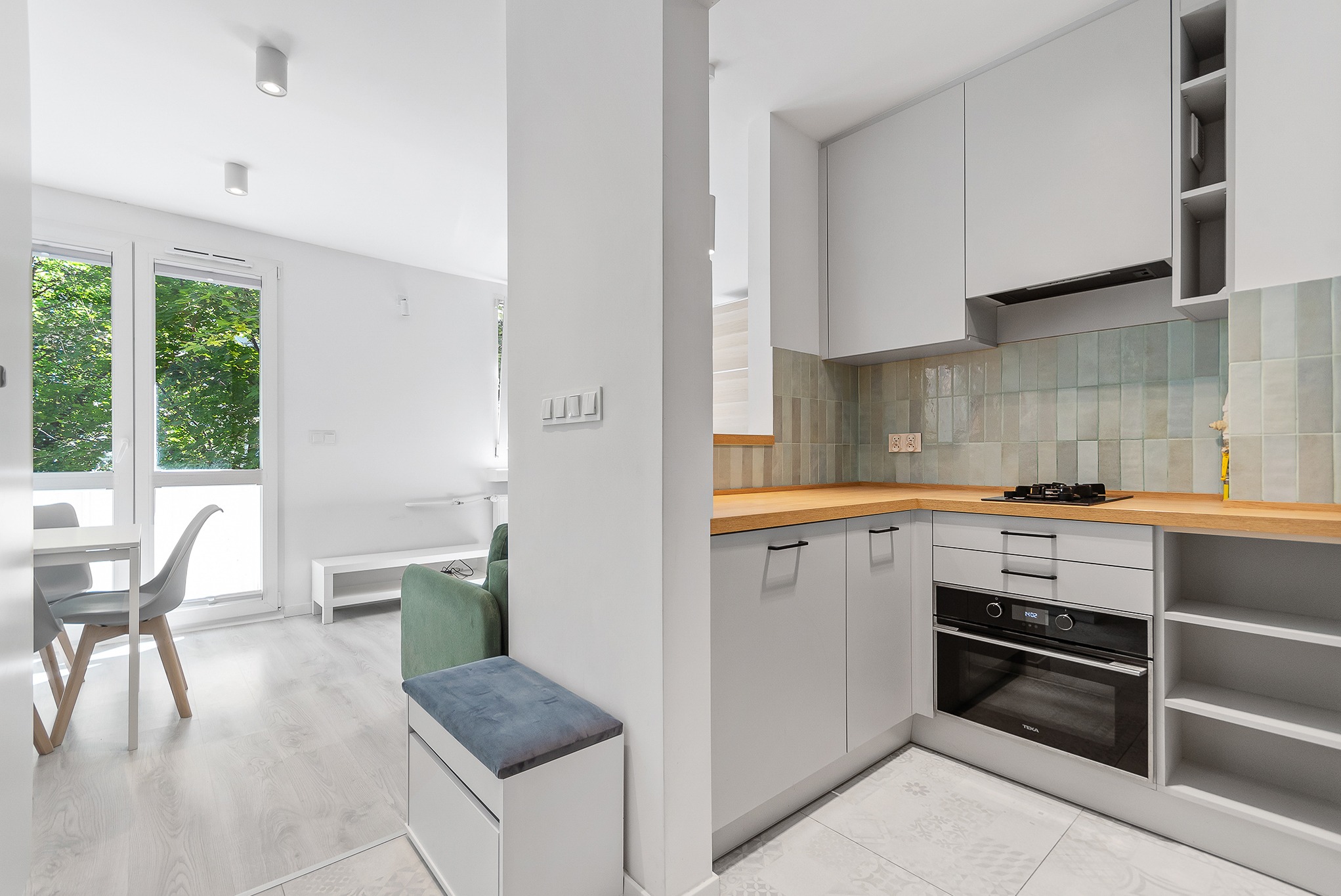 Jasne, minimalistyczne wnętrze z kuchnią w odcieniach szarości, drewnianym blatem i turkusowymi płytkami na ścianie, widok na salon z zieloną sofą i dużym oknem z widokiem na drzewa.