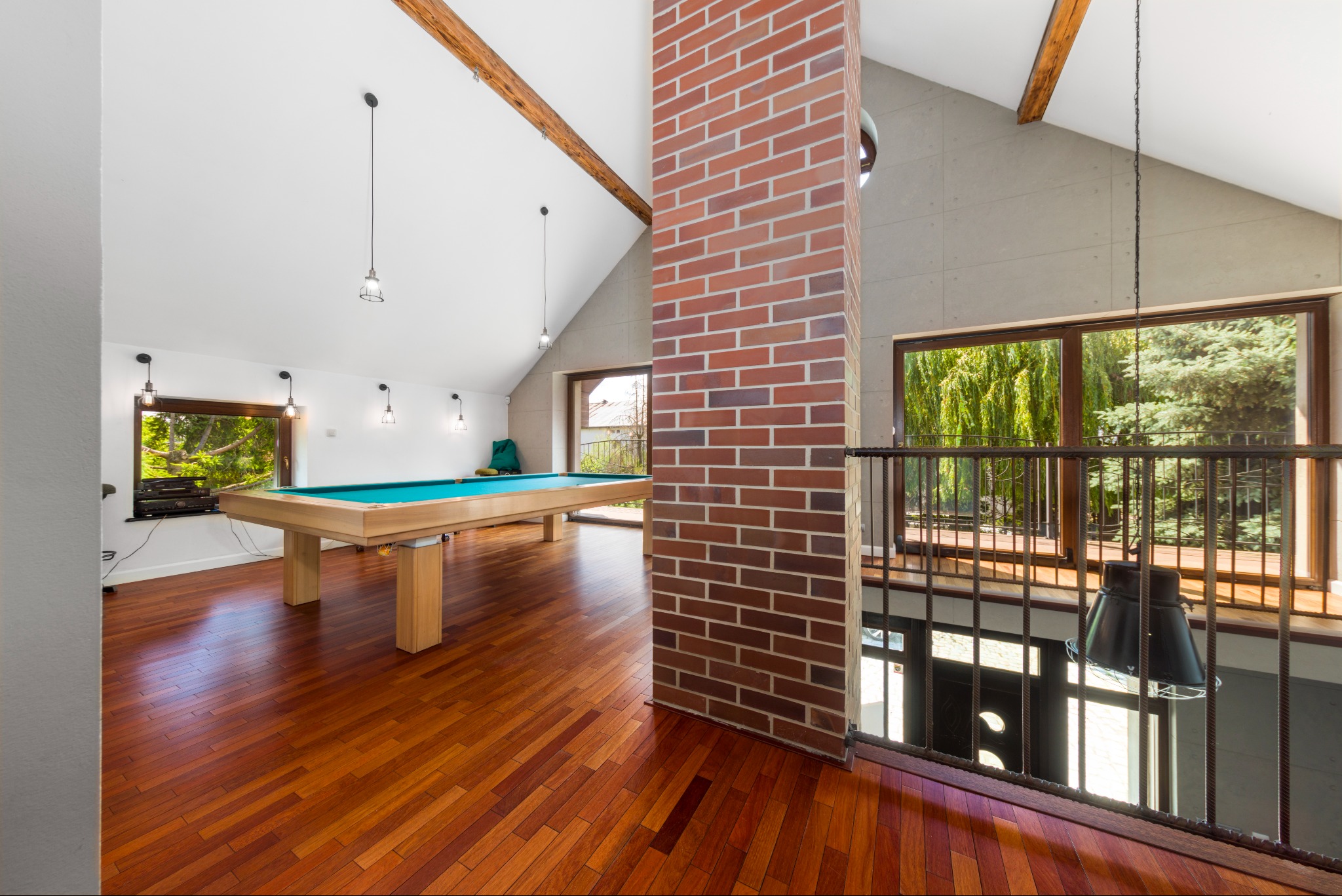 Przestronny salon z parkietem, stołem bilardowym pod skosem dachu i ceglanym kominem, widok z antresoli z czarną lampą.