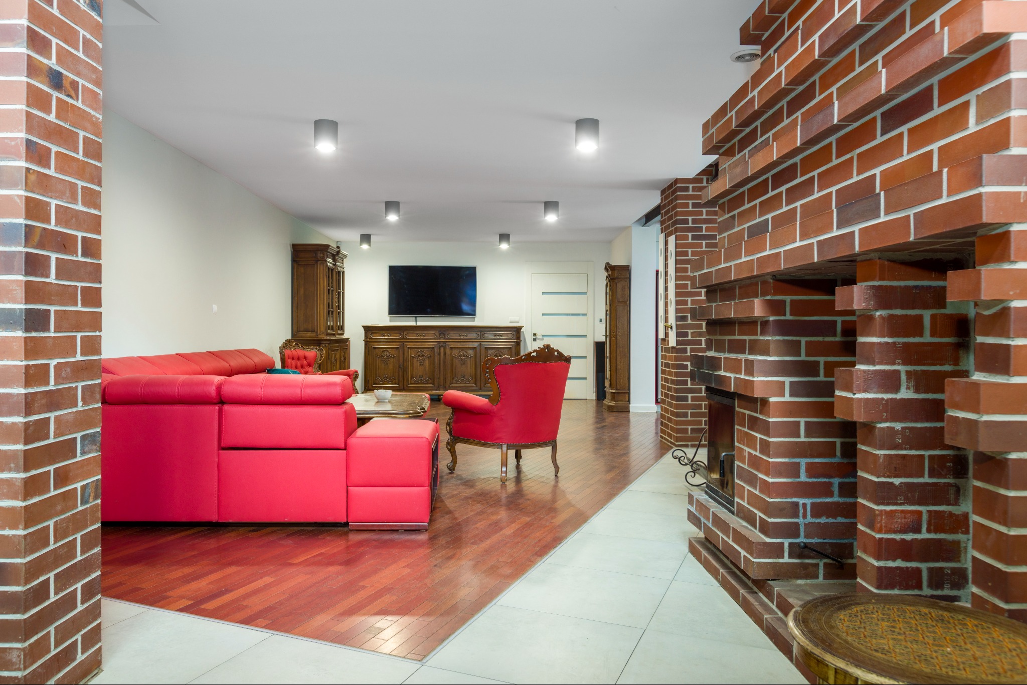 Jasne wnętrze salonu z ceglanym kominkiem, czerwonym narożnikiem i parkietem w odcieniach czerwieni.