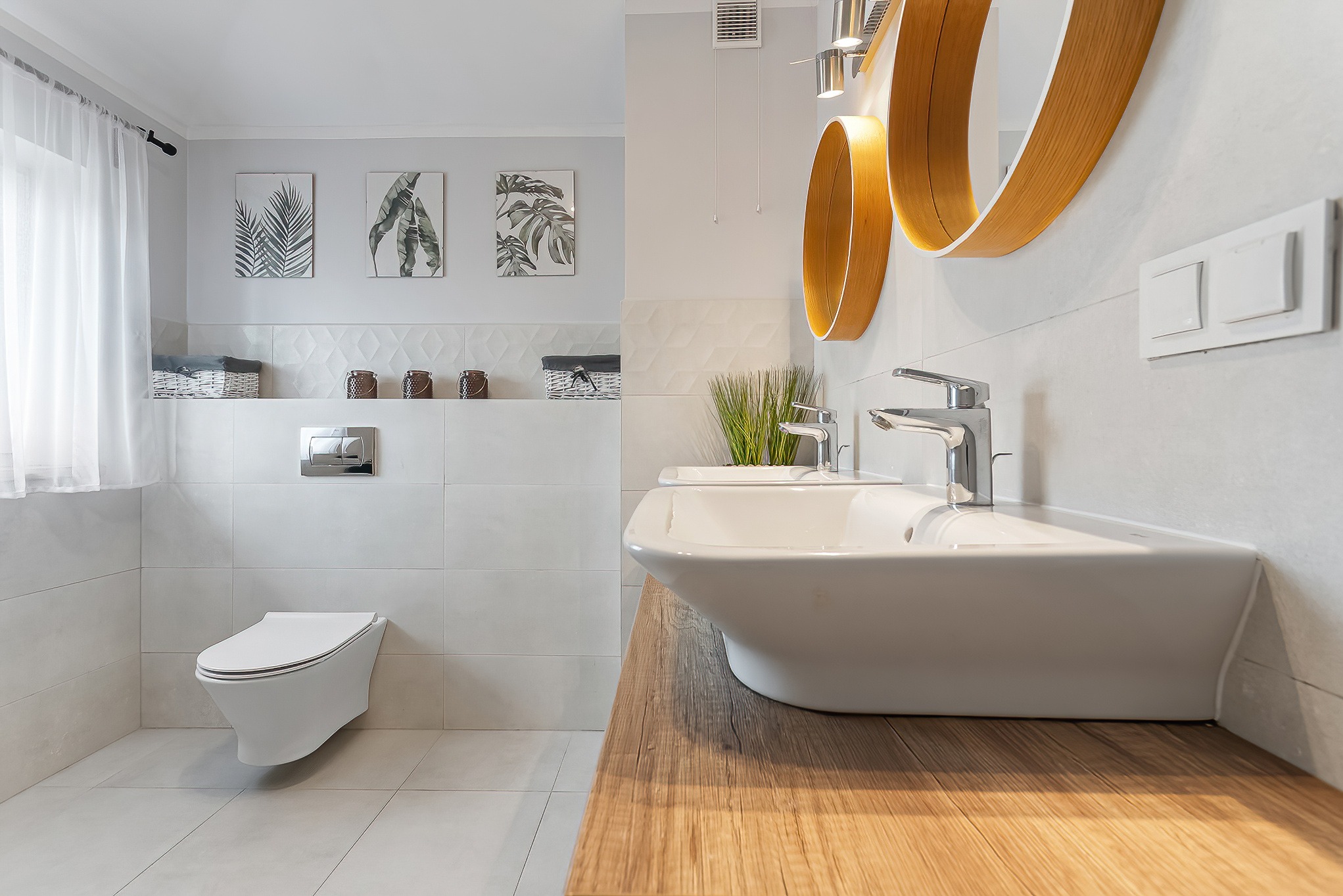 Nowoczesna łazienka w jasnych kolorach z drewnianym blatem pod umywalką, okrągłe lustra z drewnianą ramą, dekoracyjne grafiki roślinne na ścianie i wisząca toaleta