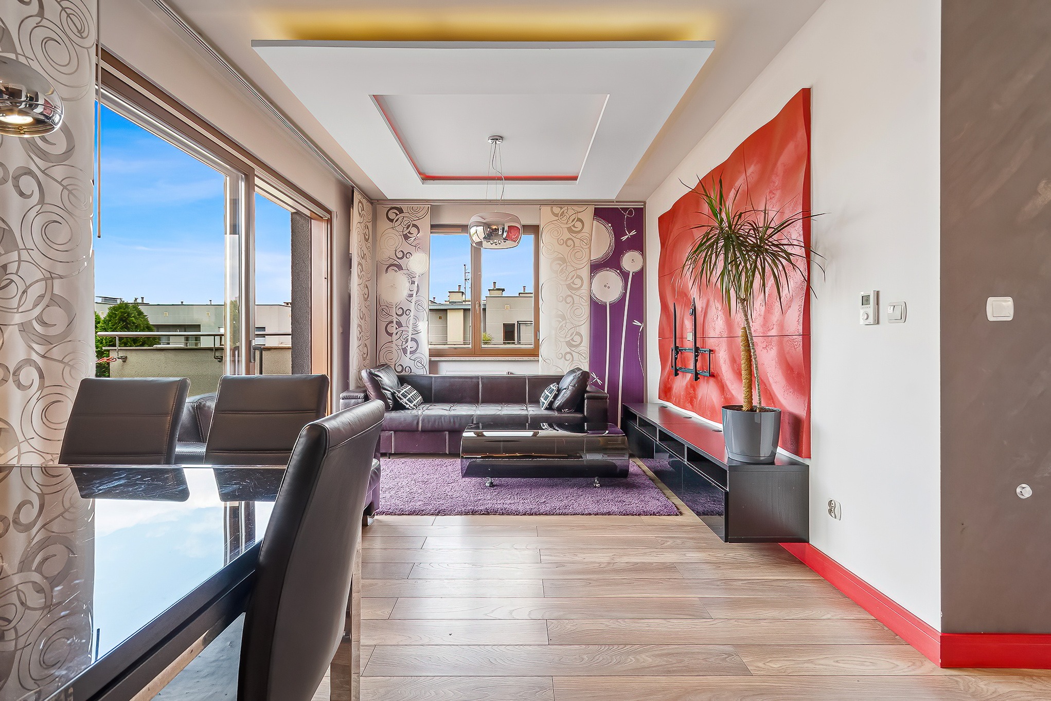 Nowoczesny salon z dużym oknem balkonowym, skórzaną sofą, fioletowym dywanem i oryginalną dekoracją ścienną w kolorze czerwonym.