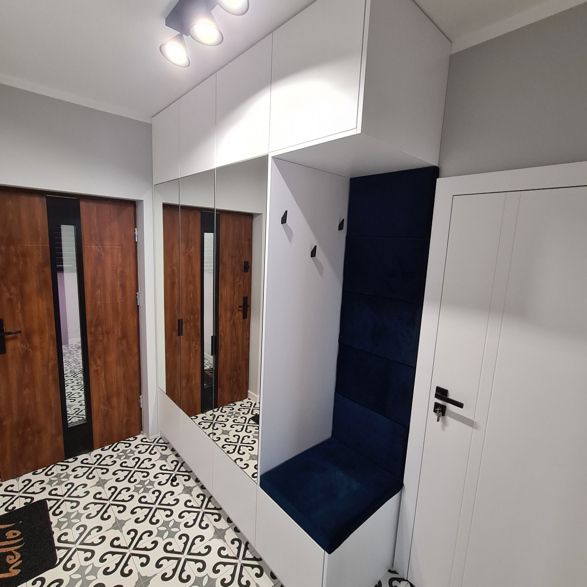 Nowoczesny przedpokój z białą szafą w zabudowie, lustrem, siedziskiem z granatową tapicerką i trzema czarnymi wieszakami, wzorzysta podłoga.