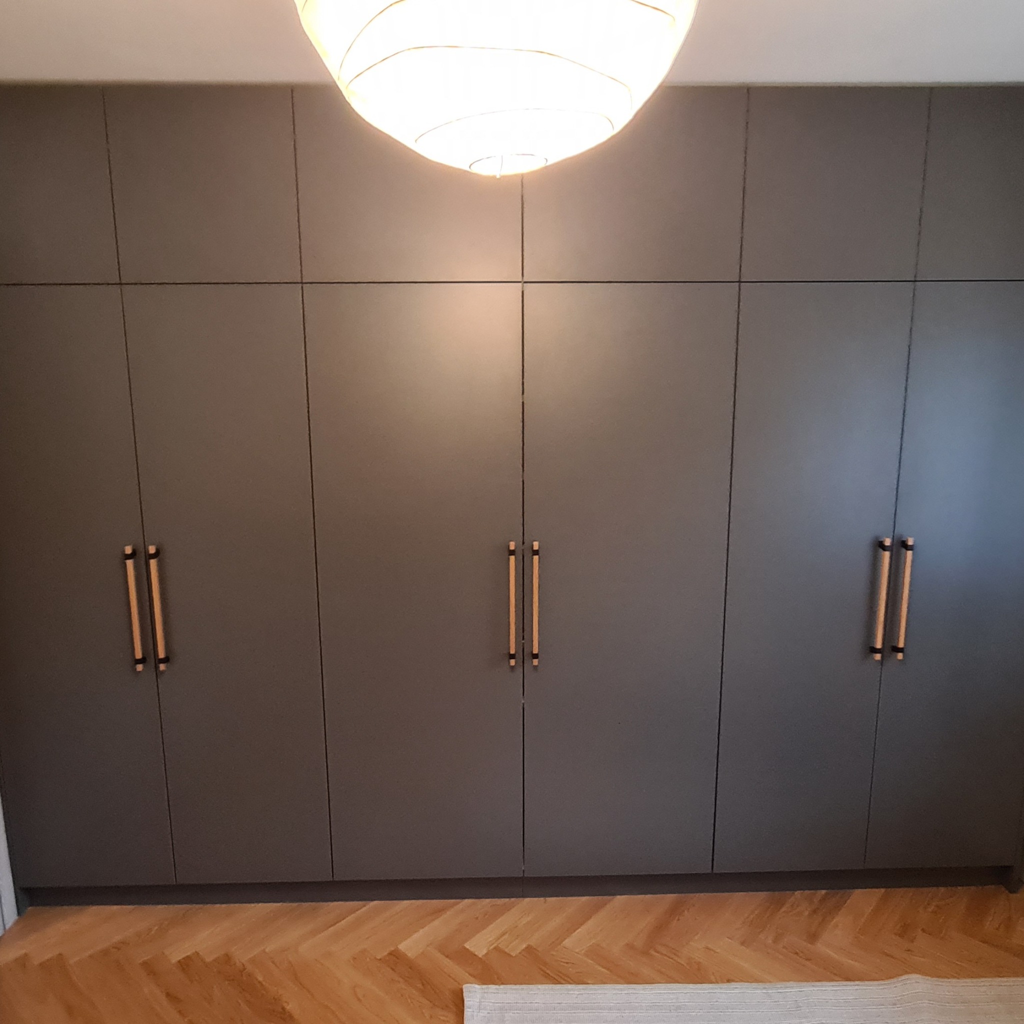 Duża, szara szafa wnękowa z drewnianymi uchwytami, widoczna na tle parkietu ułożonego w jodełkę, oświetlona przez lampę sufitową.