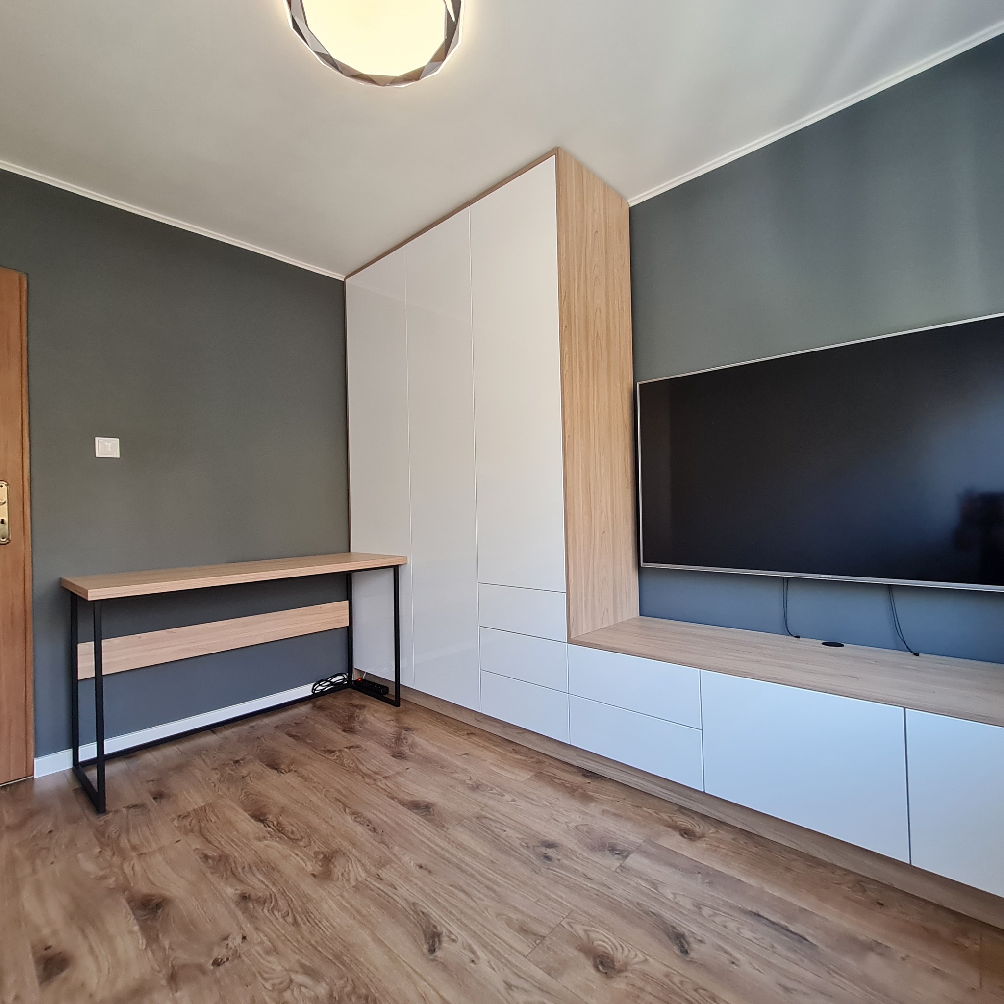 Nowoczesny pokój z białą szafą w zabudowie, drewnianymi akcentami, biurkiem z czarnymi nogami i telewizorem na ścianie w odcieniach szarości.