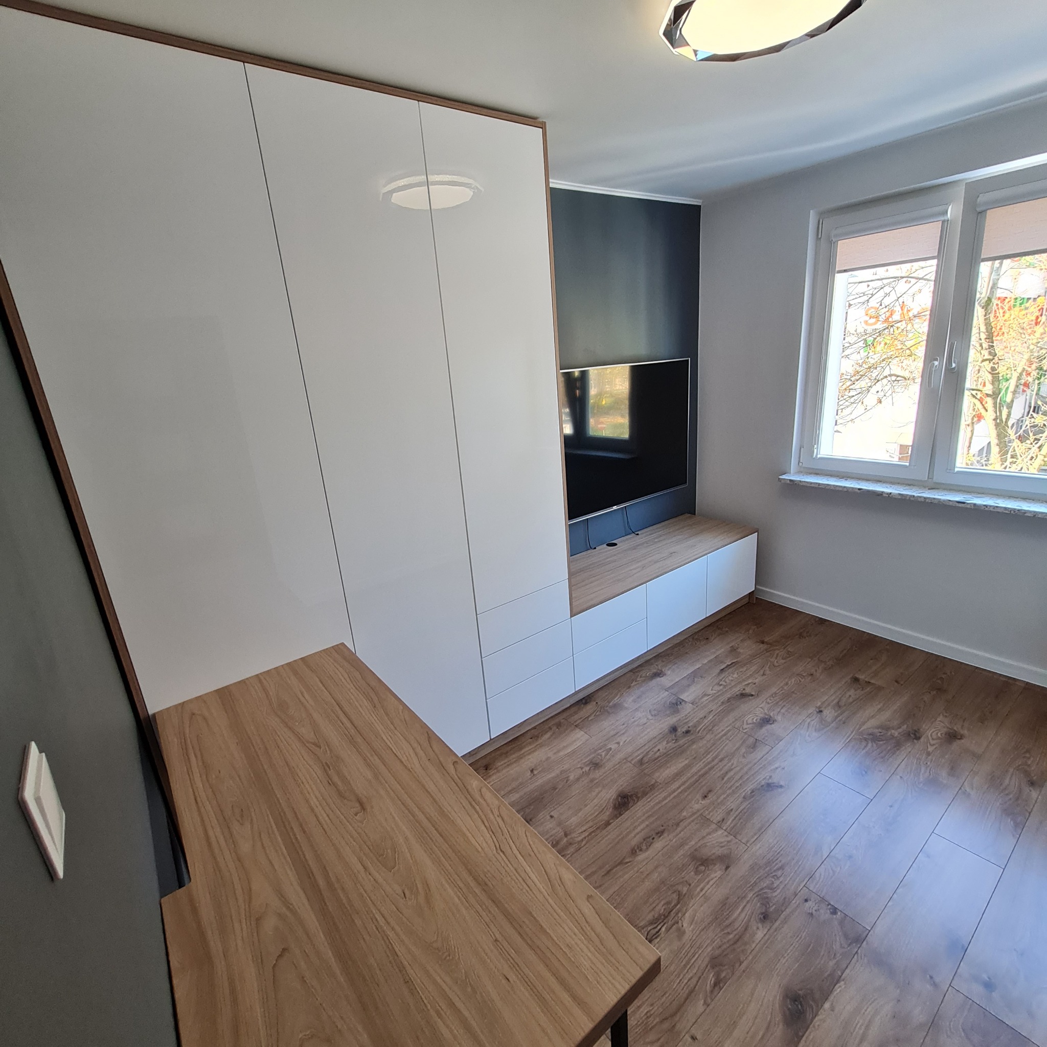 Nowoczesna szafa wnękowa z białymi frontami i drewnianymi akcentami, wbudowana w ścianę z czarnym panelem TV, drewniana podłoga, widok z boku, światło dzienne.