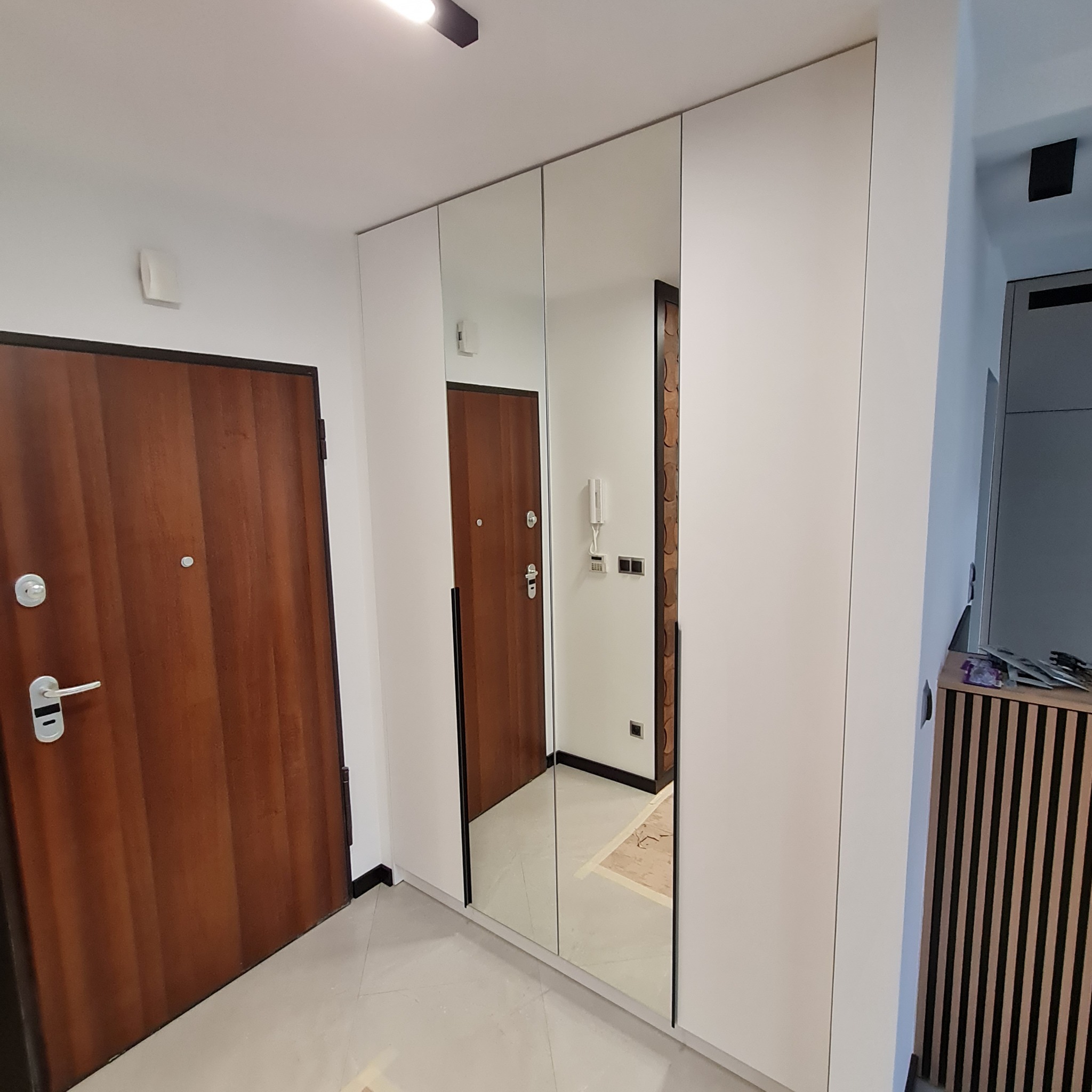 Biała szafa wnękowa z lustrzanymi drzwiami w przedpokoju, odbijająca drewniane drzwi wejściowe i fragment wnętrza mieszkania.