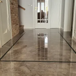 Polerowana podłoga z kamienia naturalnego w holu, z ciemnymi żyłkami i obramowaniem, przechodząca w drewniany parkiet, widok na białe drzwi z szybami.