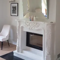 Biały, zdobiony kominek z wkładem, zwieńczony lustrem i świecami, na tle jasnych ścian i podłogi z czarnego marmuru.