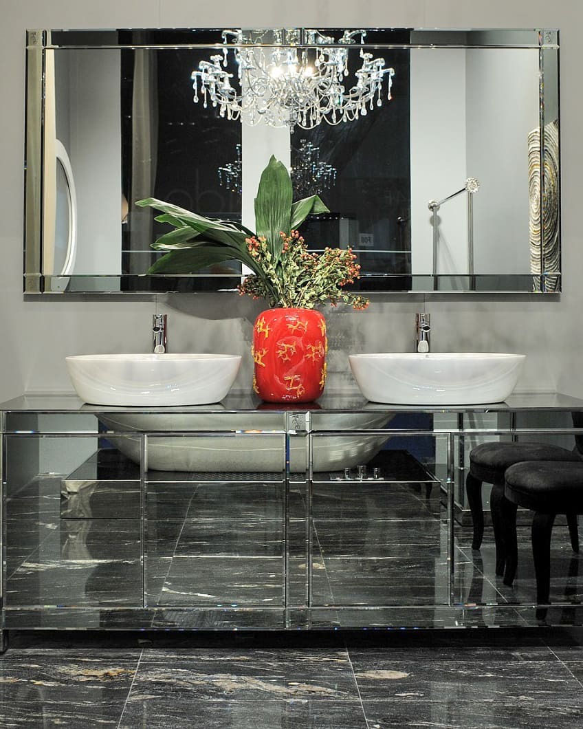 Łazienka z blatem z ciemnego kamienia, dwoma białymi umywalkami, dużym lustrem z kryształowym żyrandolem odbijającym się w nim i czerwonym wazonem z zielonymi liśćmi i czerwonymi owocami.