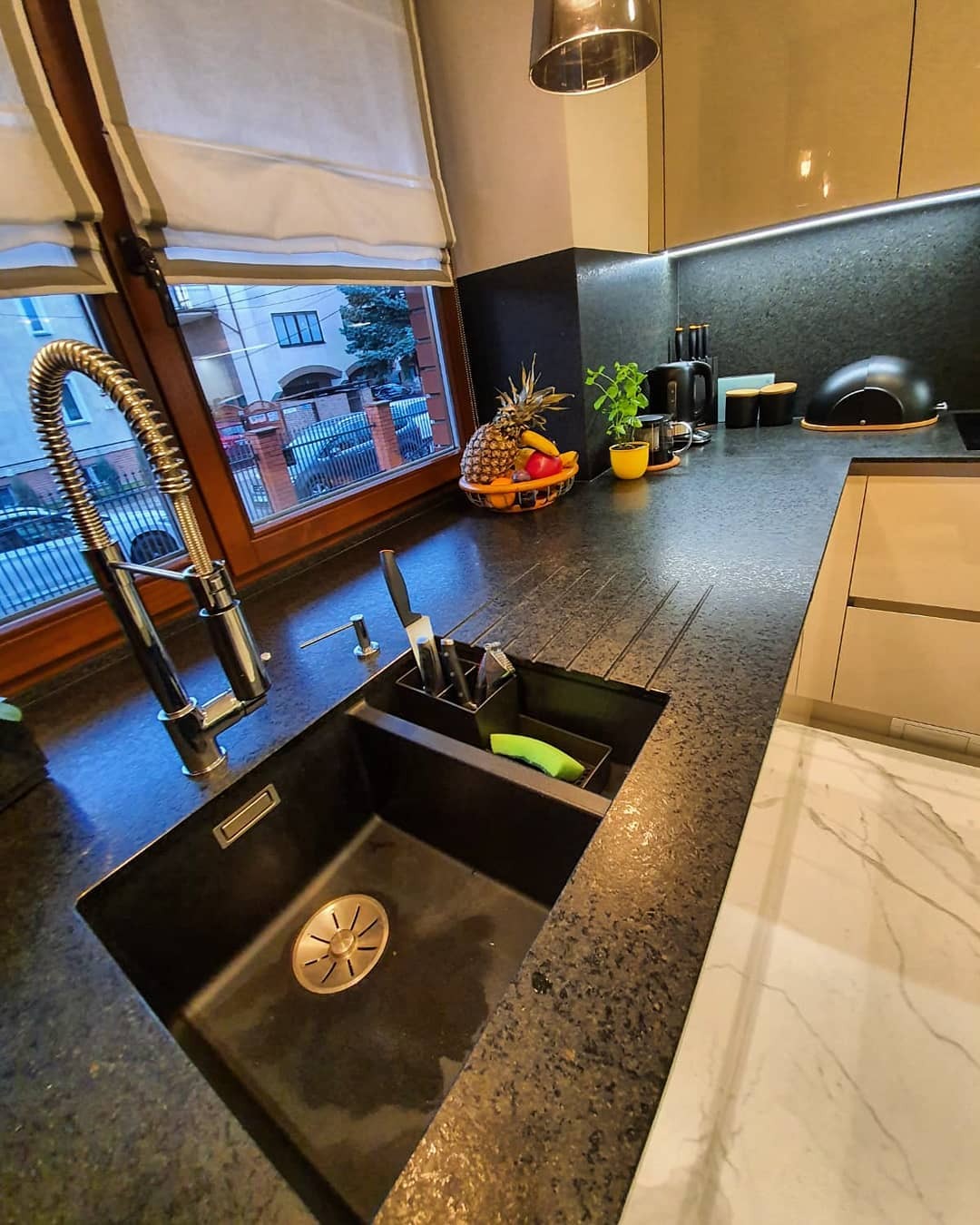 Czarny, teksturowany blat kuchenny z wbudowanym zlewozmywakiem i baterią w nowoczesnej kuchni. Na blacie widoczny koszyk z owocami, przyprawy i akcesoria kuchenne, a w tle okno z widokiem na ulicę.