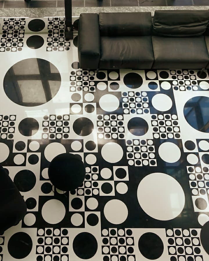 Wzorzysta posadzka z czarno-białych okręgów i kwadratów, częściowo zasłonięta przez czarną sofę modułową i fragment czarnego krzesła.