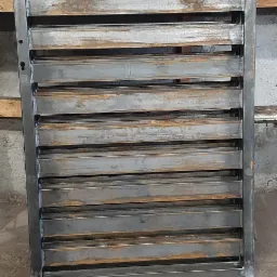 Surowa, metalowa konstrukcja z poziomymi lamelami, prawdopodobnie element ogrodzenia lub bramy, widoczne ślady spawania i obróbki, na tle betonowej ściany i podłogi.
