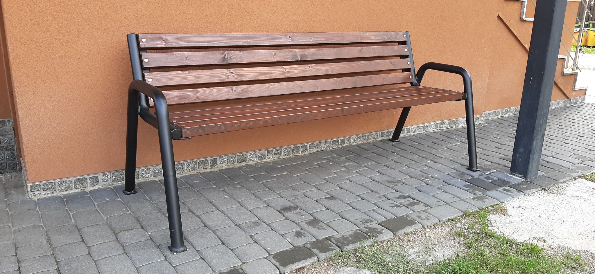Nowoczesna ławka parkowa z brązowego drewna i czarnego metalu, ustawiona na szarej kostce brukowej przed budynkiem o pomarańczowej elewacji.