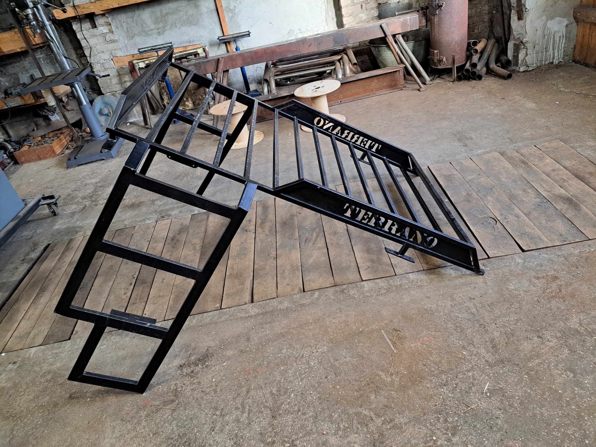 Czarne, metalowe schody z napisem TERRAZO, leżące w warsztacie na drewnianej podłodze, w tle widoczne narzędzia i materiały.