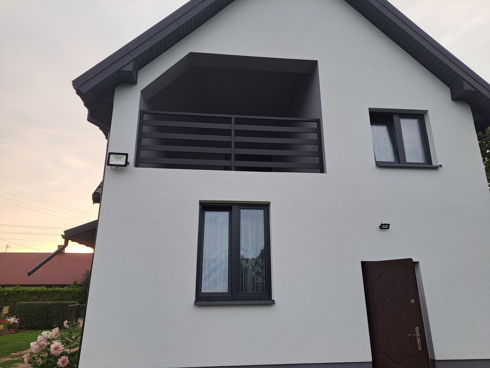 Nowoczesny dom jednorodzinny z balkonem zabezpieczonym czarną, poziomą balustradą. Widoczne okna z ciemnymi ramami, oświetlenie zewnętrzne oraz częściowo otwarte drzwi wejściowe.