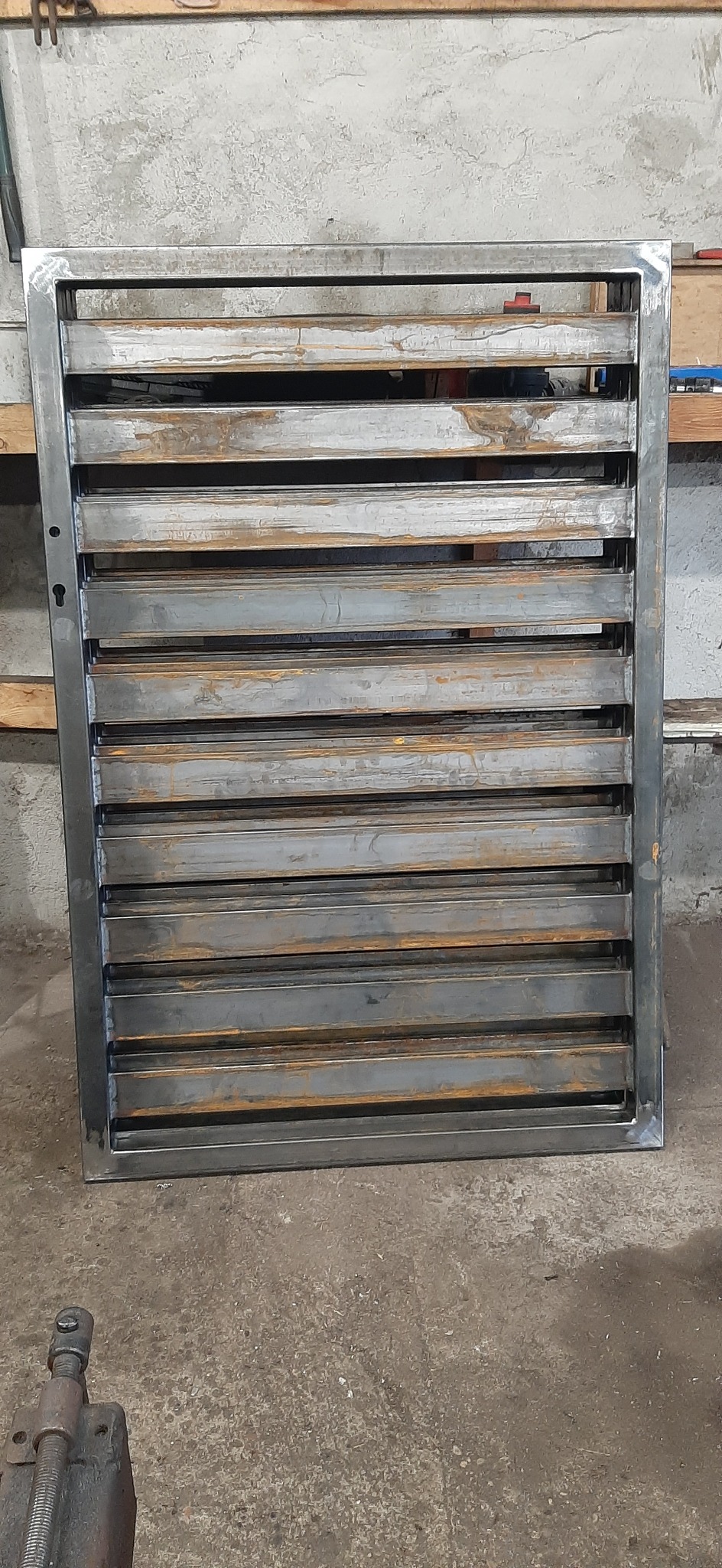 Surowa, metalowa konstrukcja z poziomymi lamelami, prawdopodobnie element ogrodzenia lub kraty, widoczne ślady rdzy i niedoskonałości spoin.