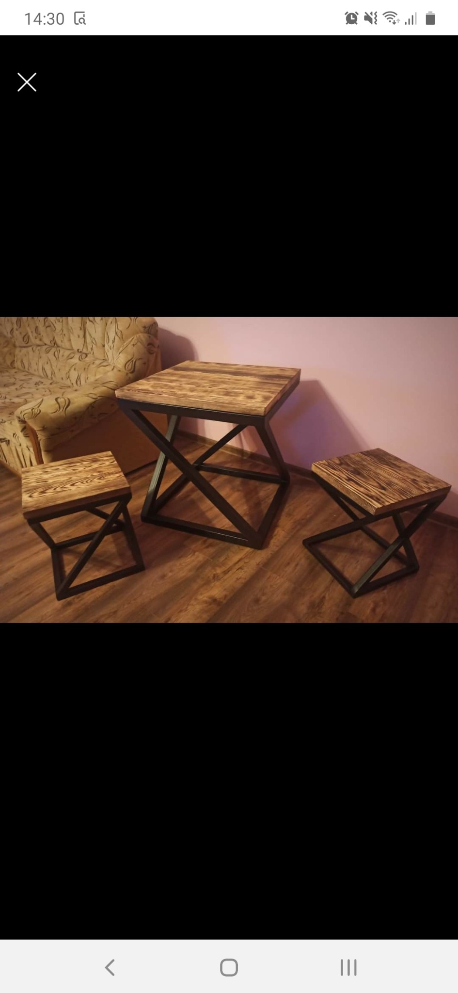 Stół i dwa stołki z drewnianymi blatami o wyraźnym usłojeniu i czarnymi, metalowymi nogami o geometrycznym kształcie X, ustawione na drewnianej podłodze w pokoju z beżową kanapą i różowymi ścianami.