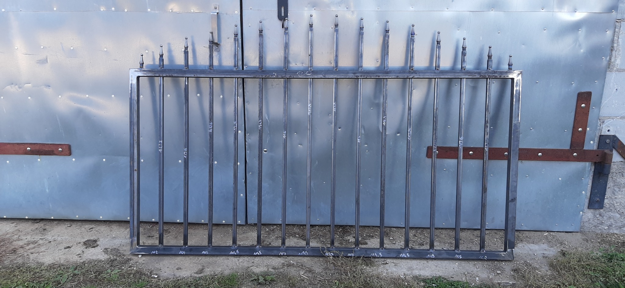 Surowy, metalowy fragment ogrodzenia z pionowymi prętami i ozdobnymi zakończeniami, oparty o blaszane, garażowe wrota z widocznymi śladami spawania i numerami identyfikacyjnymi naniesionymi na pręty.