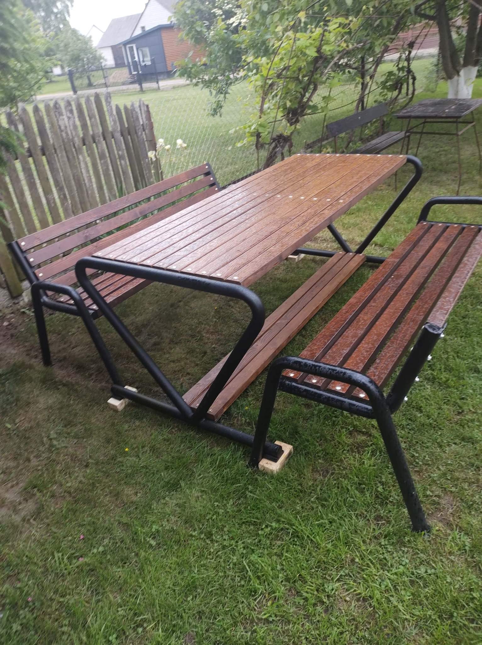 Drewniany stół piknikowy z ławkami na metalowym stelażu, stojący na trawie w ogrodzie, widoczne krople wody po deszczu.