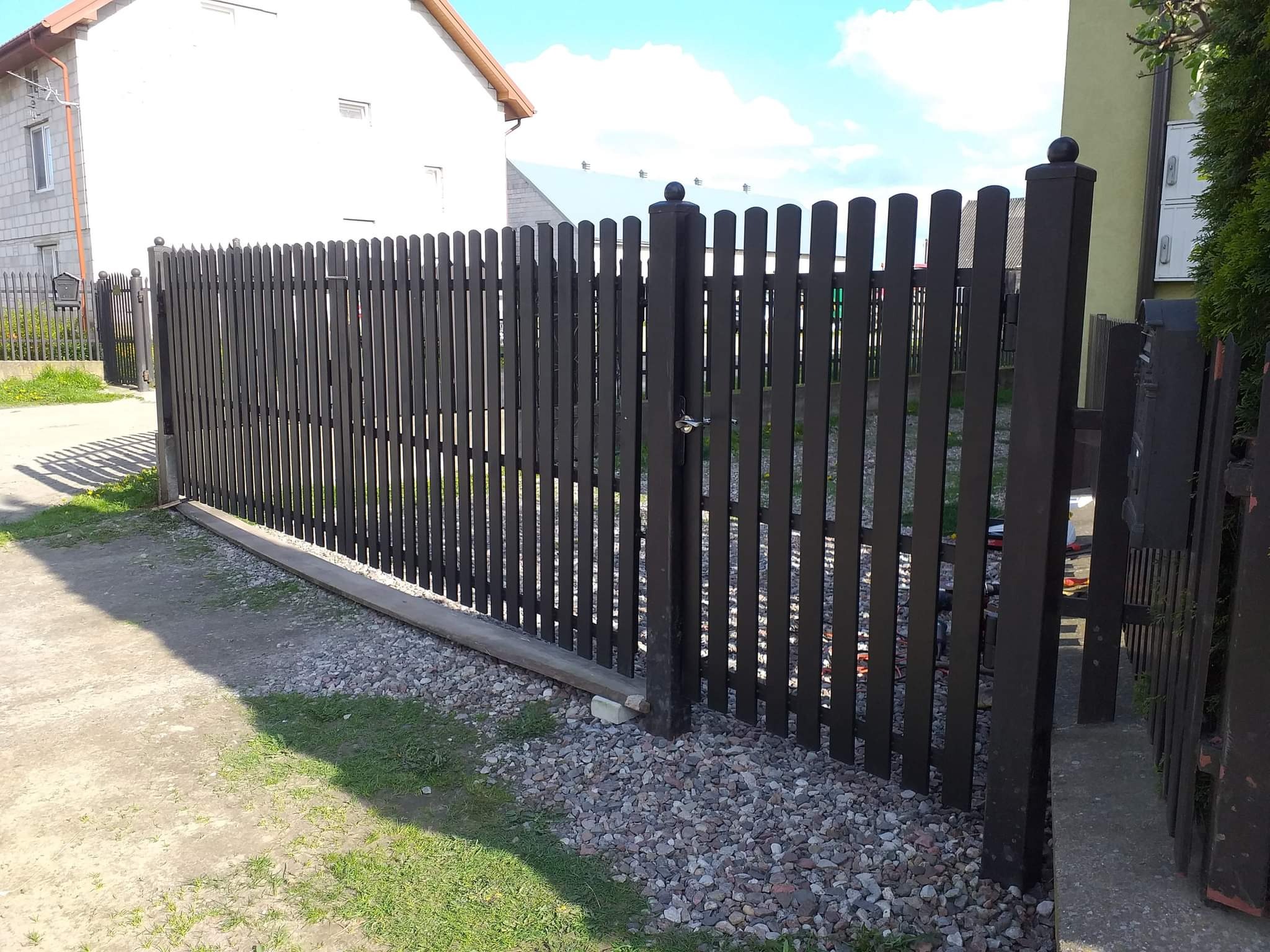 Ciemne, pionowe ogrodzenie z metalowych paneli z okrągłymi zakończeniami słupków, z widoczną furtką, na tle jasnego domu i niebieskiego nieba.