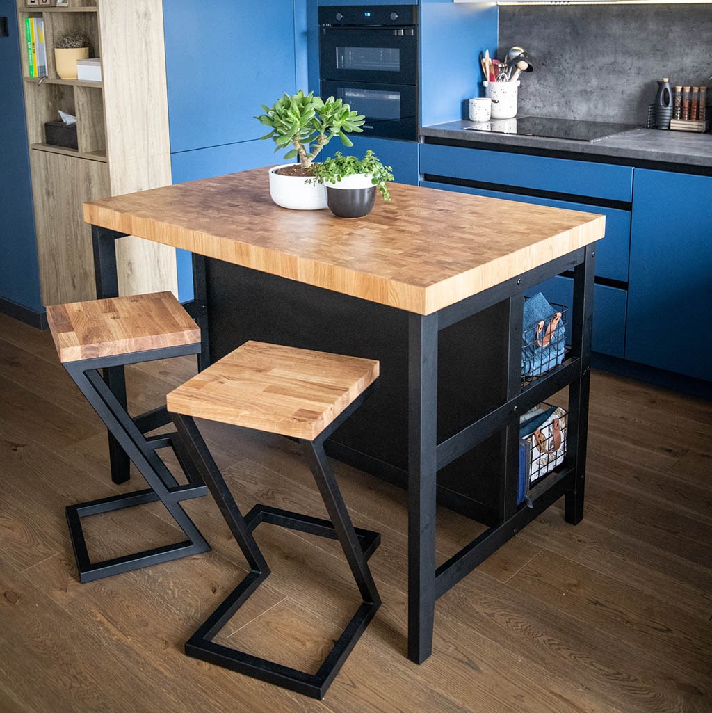 Wyspa kuchenna z blatem z klejonki dębowej, czarną metalową konstrukcją i dwoma hokerami o geometrycznych nogach, uzupełniona roślinami doniczkowymi na tle błękitnych szafek kuchennych.