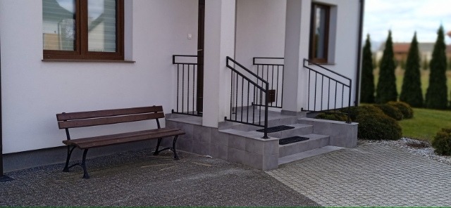 Zewnętrzne schody wejściowe do domu z czarnymi, metalowymi balustradami, wyłożone szarymi płytkami, z drewnianą ławką obok.